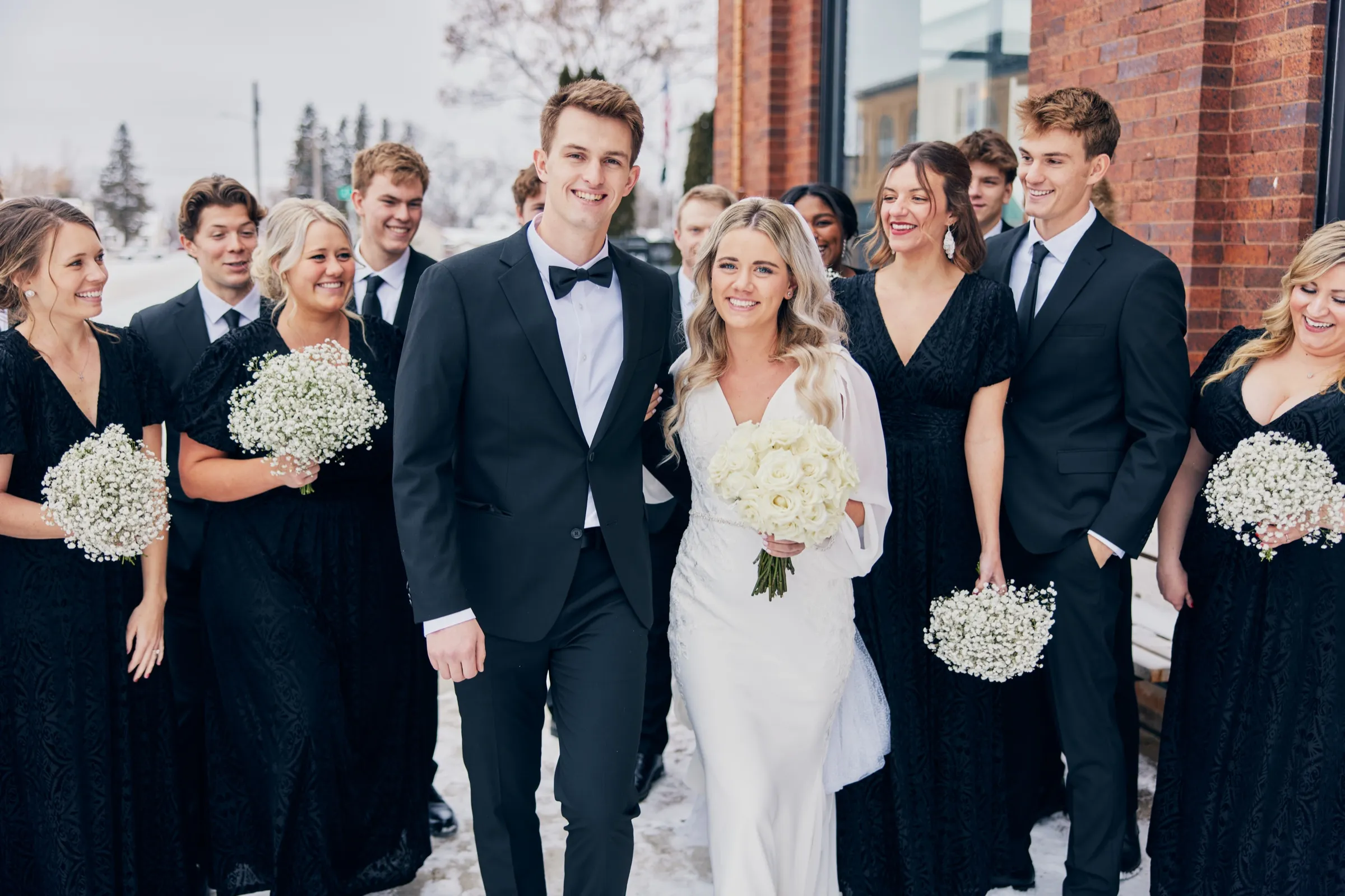 Madeline and Sam walk forward flanked by their bridal party in black, holding white baby's breath, along a snowy sidewalk beside a brick building — Tim Larsen Photography, Brainerd Lakes MN