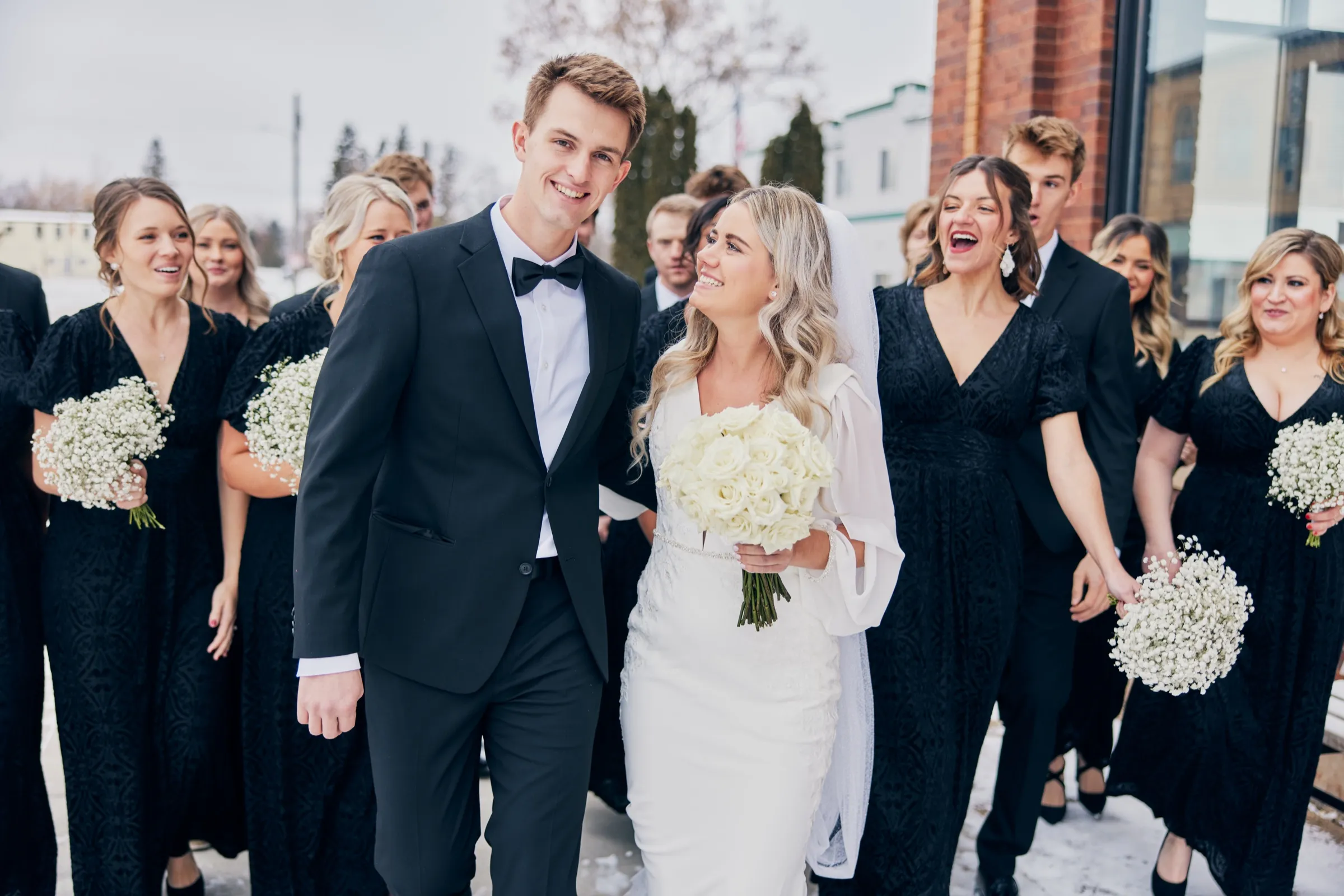 Madeline and Sam walk forward laughing with their wedding party in black formalwear, holding white baby's-breath bouquets, beside a brick building in winter — Tim Larsen Photography, Brainerd Lakes MN