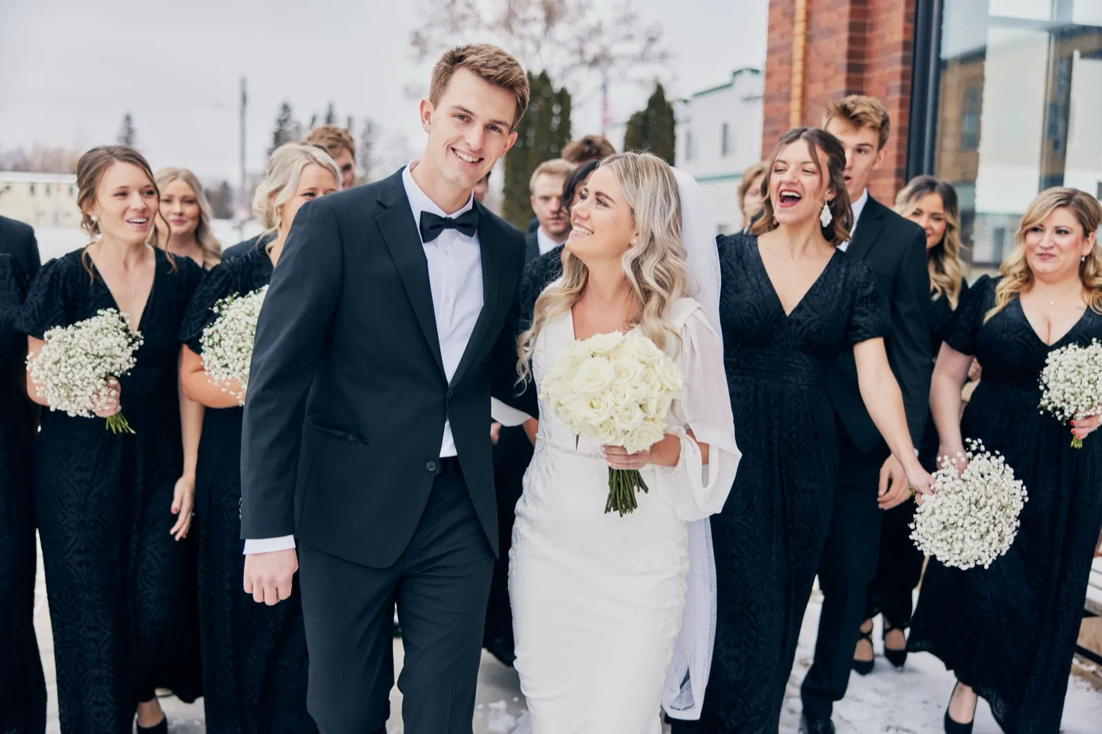 The bride and groom walk forward laughing alongside their bridal party in black formalwear, holding white baby's-breath bouquets, beside a brick building in winter. — Tim Larsen Photography, Brainerd Lakes MN