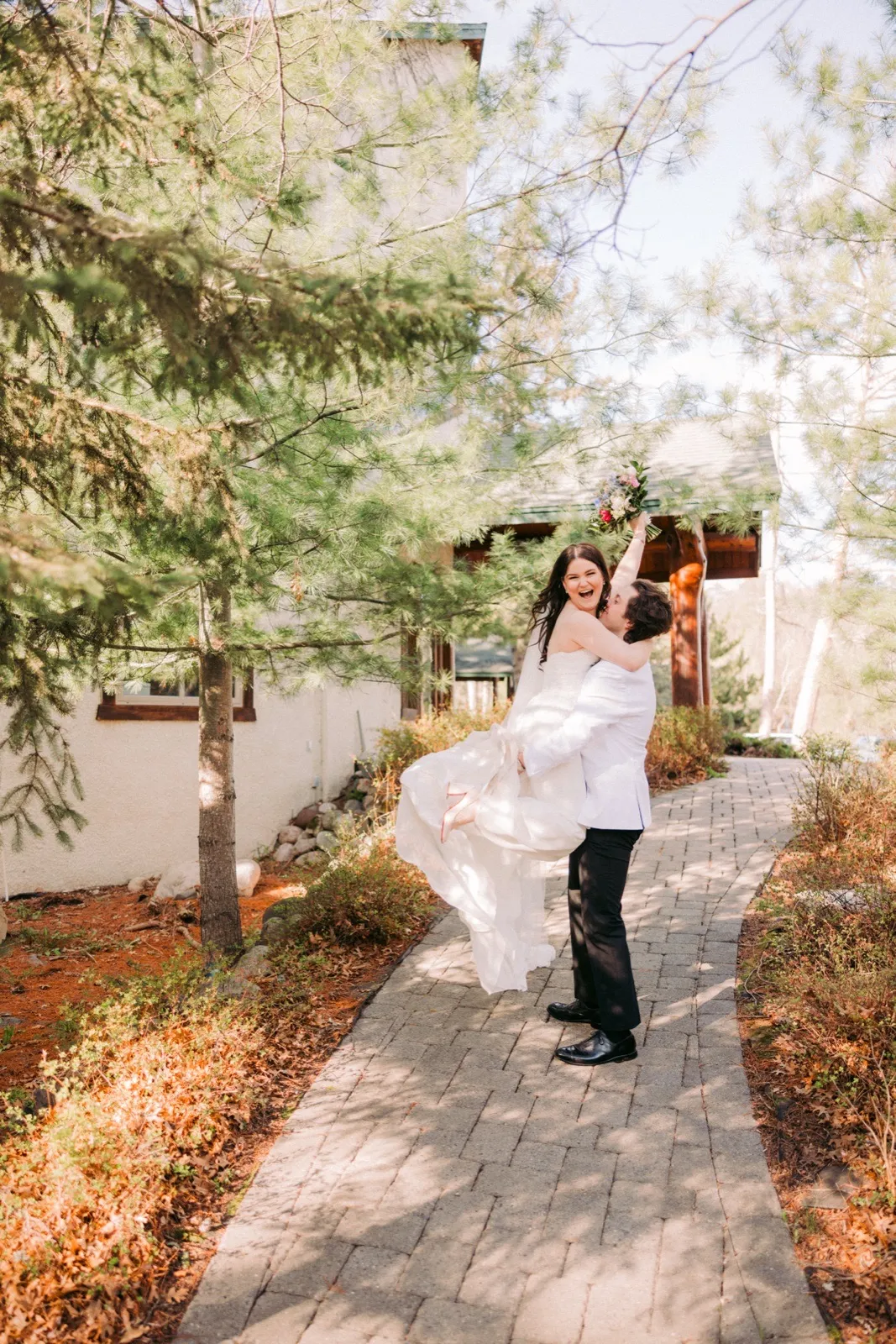 The groom lifts the bride off the ground as she raises her bouquet and laughs on a paver pathway outside Manhattan Beach Lodge — Tim Larsen Photography, Brainerd Lakes MN