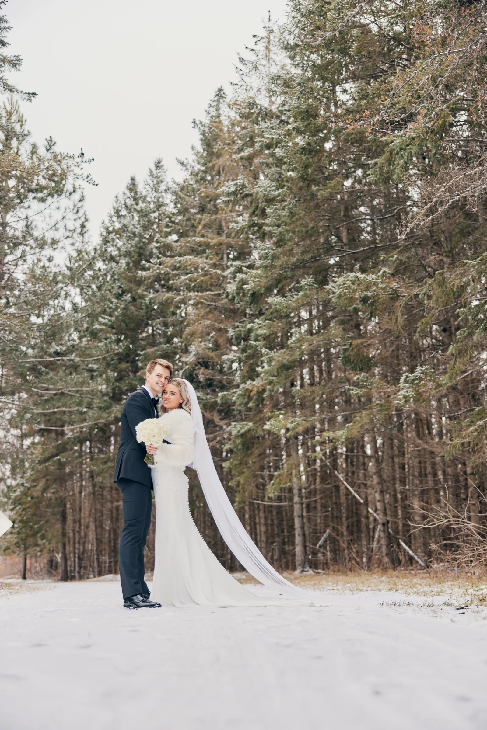 Madeline and Sam stand close together in a snowy clearing surrounded by tall pines, her long veil trailing across the snow — Tim Larsen Photography, Brainerd Lakes MN
