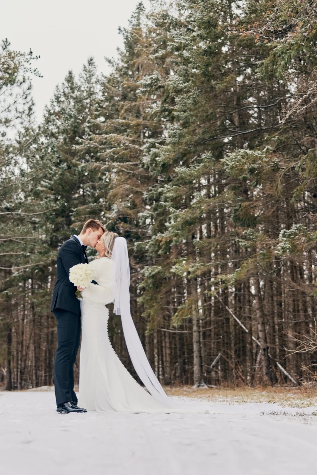 The bride and groom kiss in a snowy clearing while her long veil sweeps across the snow in front of a tall pine forest. — Tim Larsen Photography, Brainerd Lakes MN