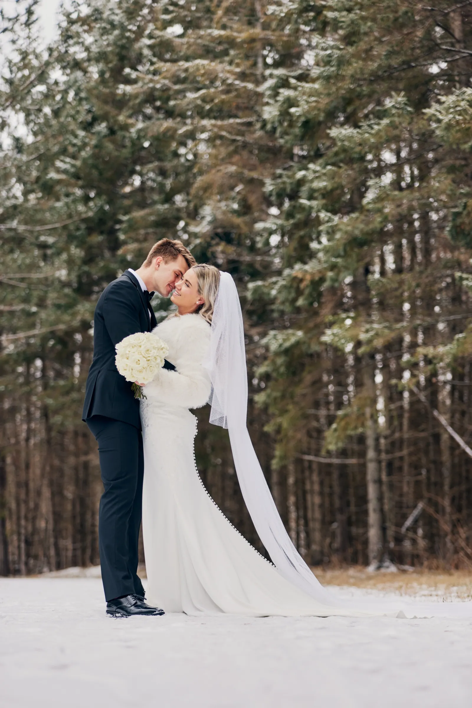 Madeline and Sam stand forehead to forehead in a snowy clearing, her veil and gown trailing across the snow against a backdrop of evergreens — Tim Larsen Photography, Brainerd Lakes MN