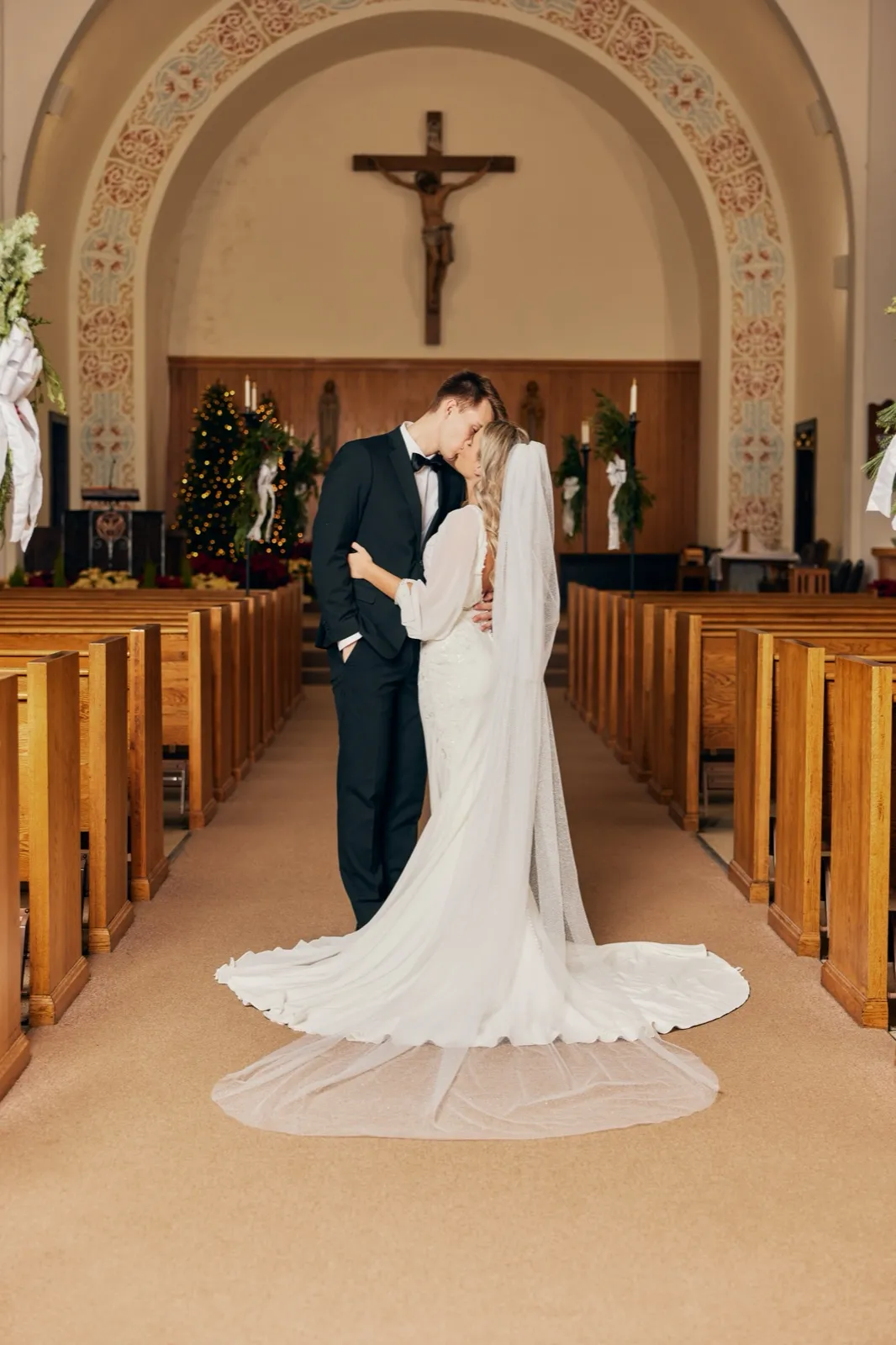 The bride and groom kiss in the center aisle of a Catholic church beneath a crucifix and decorated archway, her long veil and gown train fanned out behind her on the floor. — Tim Larsen Photography, Brainerd Lakes MN