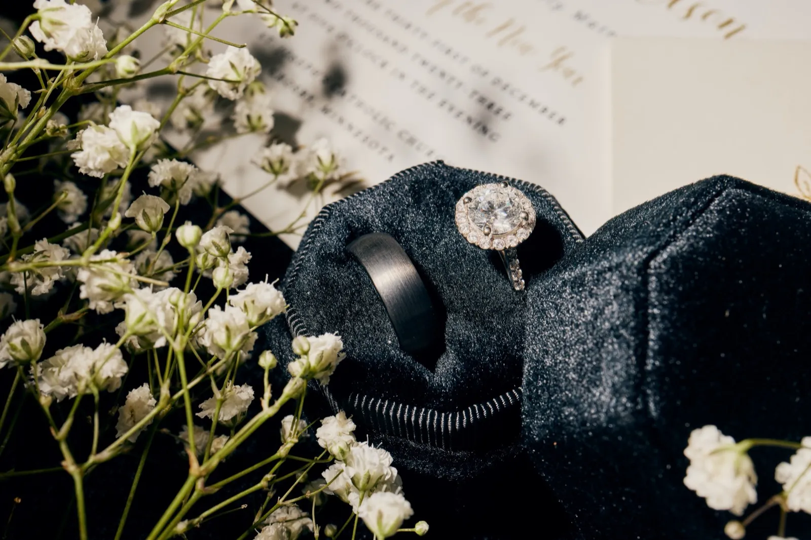 An engagement ring with a halo diamond and a brushed black wedding band rest in a black velvet box atop a printed wedding invitation, surrounded by sprigs of baby's breath. — Tim Larsen Photography, Brainerd Lakes MN