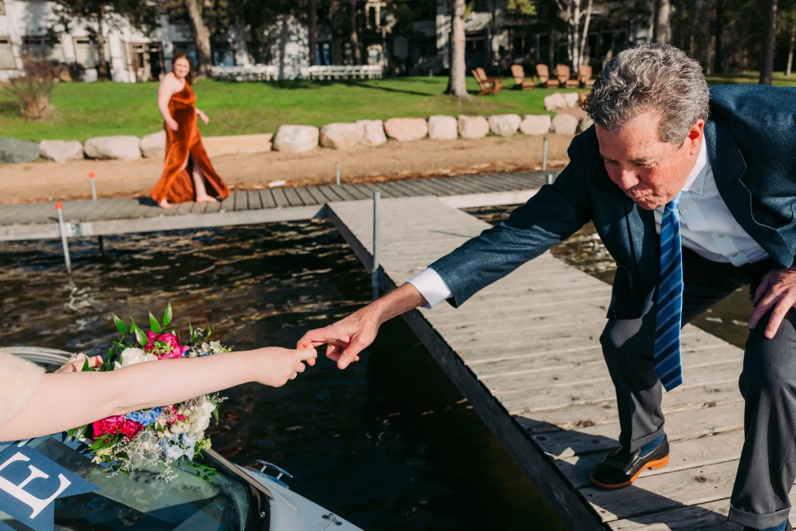 A man in a navy suit and striped tie leans across a gap in the dock to reach the bride's outstretched fingertips while a bridesmaid in a rust-colored gown walks along the shore behind them — Tim Larsen Photography, Brainerd Lakes MN