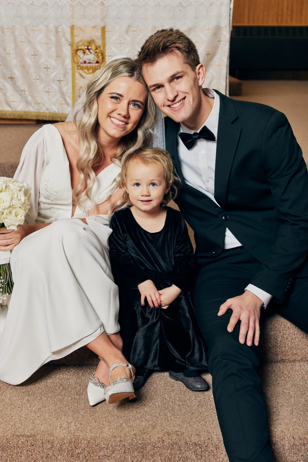 The bride and groom sit on altar steps with a small child in a black velvet dress between them, all smiling at the camera. — Tim Larsen Photography, Brainerd Lakes MN