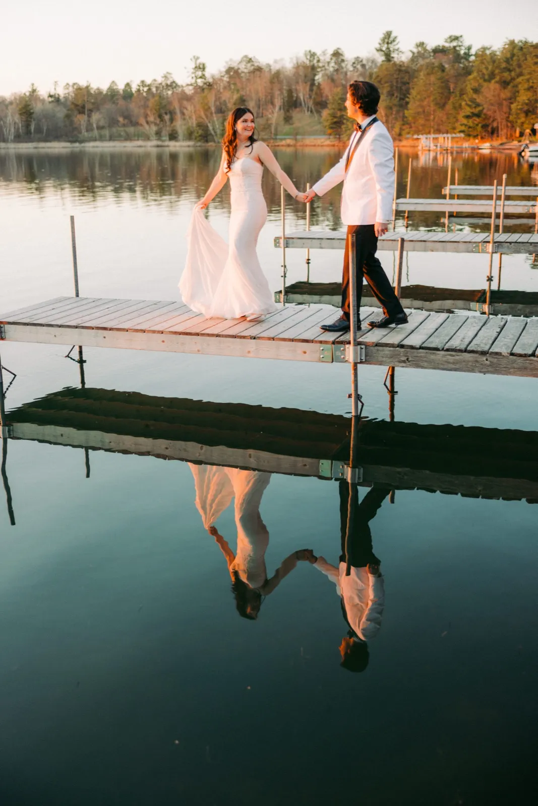 The bride and groom walk hand in hand along a wooden dock at golden hour, their figures mirrored in the still lake water below — Tim Larsen Photography, Brainerd Lakes MN