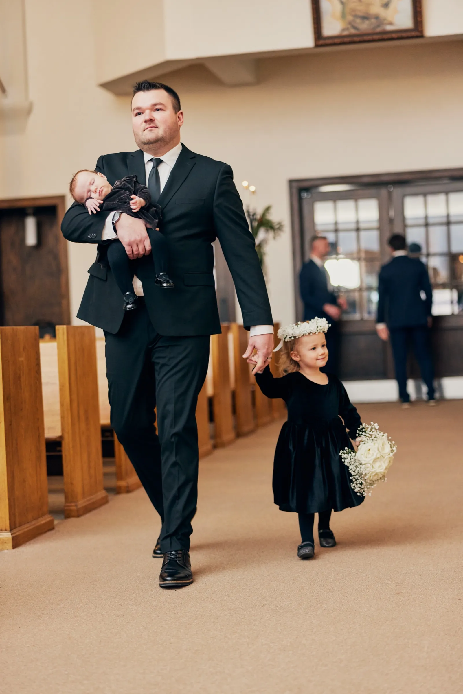 A man in a black suit carries a sleeping child down the church aisle while holding the hand of a small flower girl in a black velvet dress and floral crown — Tim Larsen Photography, Brainerd Lakes MN