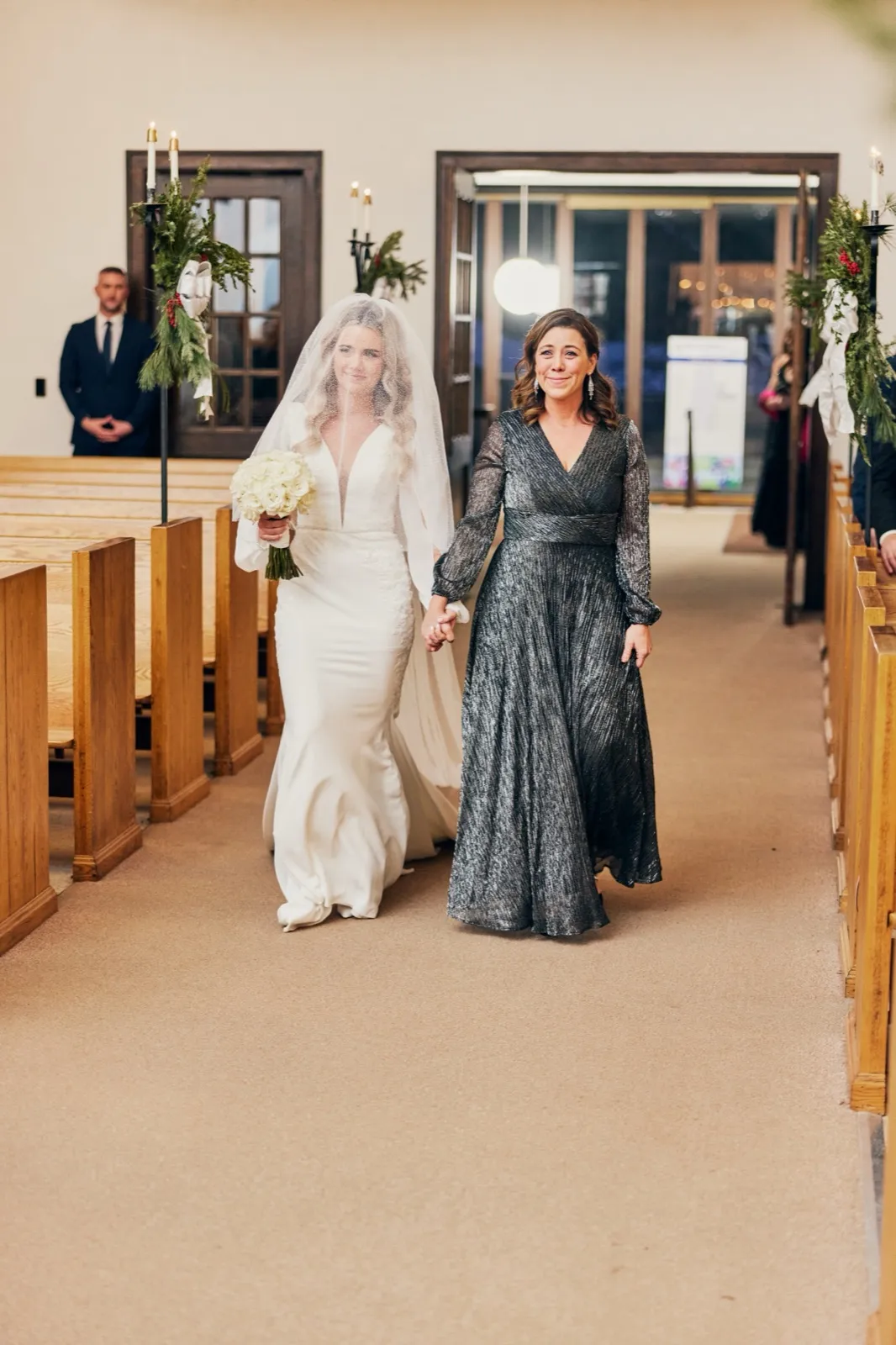 The bride walks down a candlelit chapel aisle holding her mother's hand, wearing a fitted gown and long veil while carrying a white bouquet. — Tim Larsen Photography, Brainerd Lakes MN