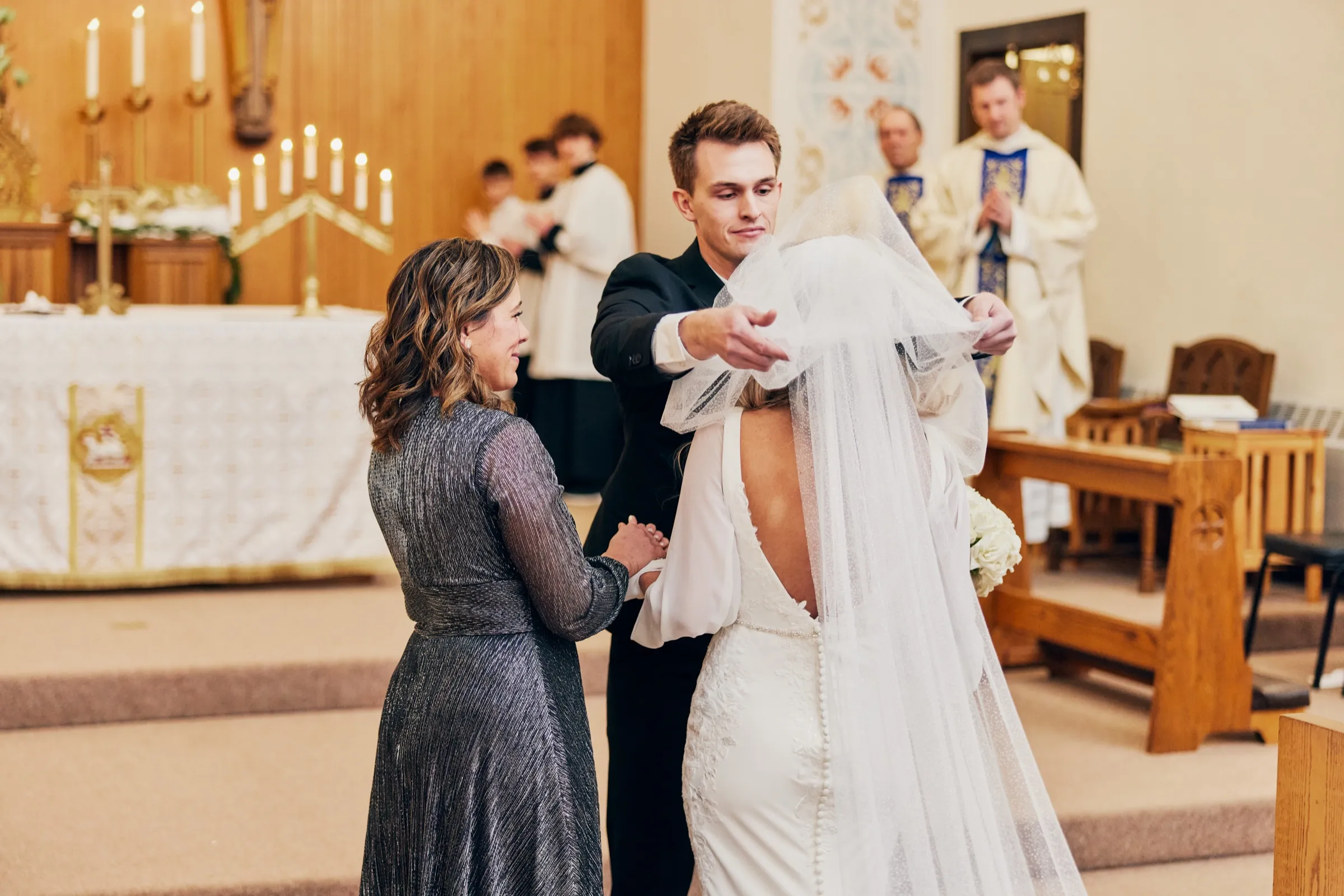 Sam carefully lifts Madeline's veil at the altar while her mother looks on with visible emotion and the priest stands behind them — Tim Larsen Photography, Brainerd Lakes MN