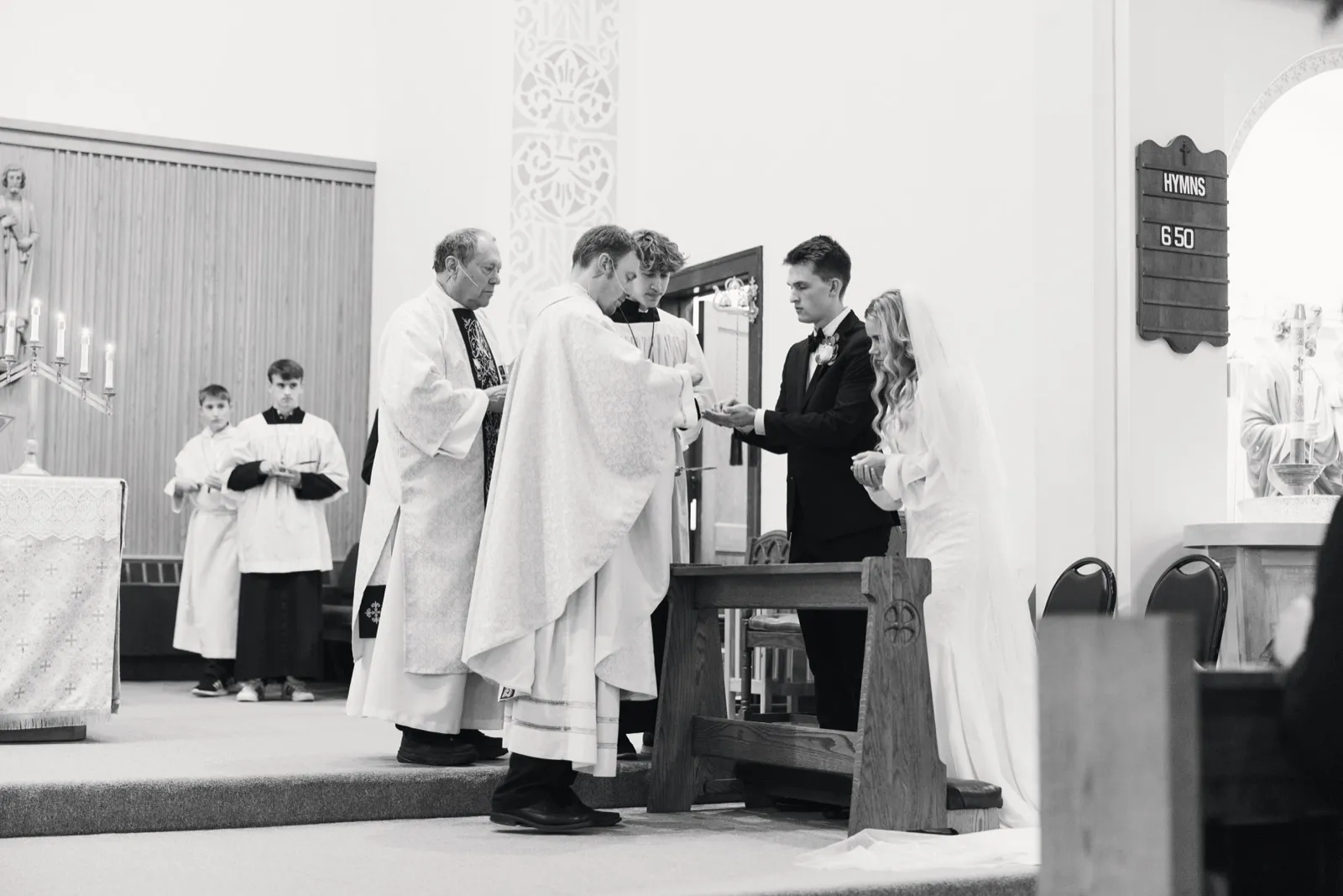 The bride in a long veil and the groom in a tuxedo exchange rings at the altar while a priest and two altar servers stand alongside them in a Catholic church. — Tim Larsen Photography, Brainerd Lakes MN