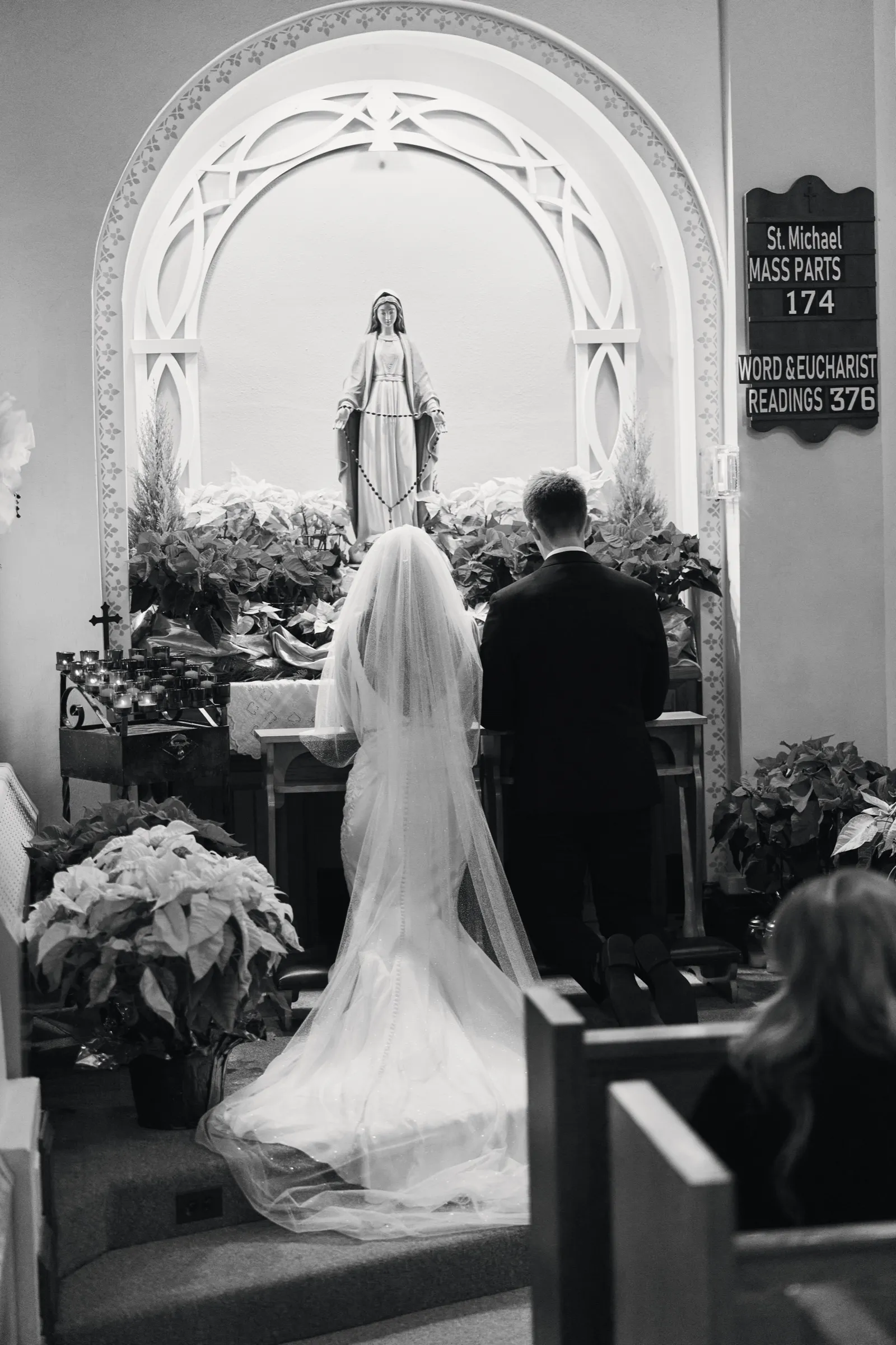 Madeline and Sam kneel together at the altar beneath an arched alcove holding a statue of Mary, surrounded by poinsettias, her veil spread across the floor — Tim Larsen Photography, Brainerd Lakes MN