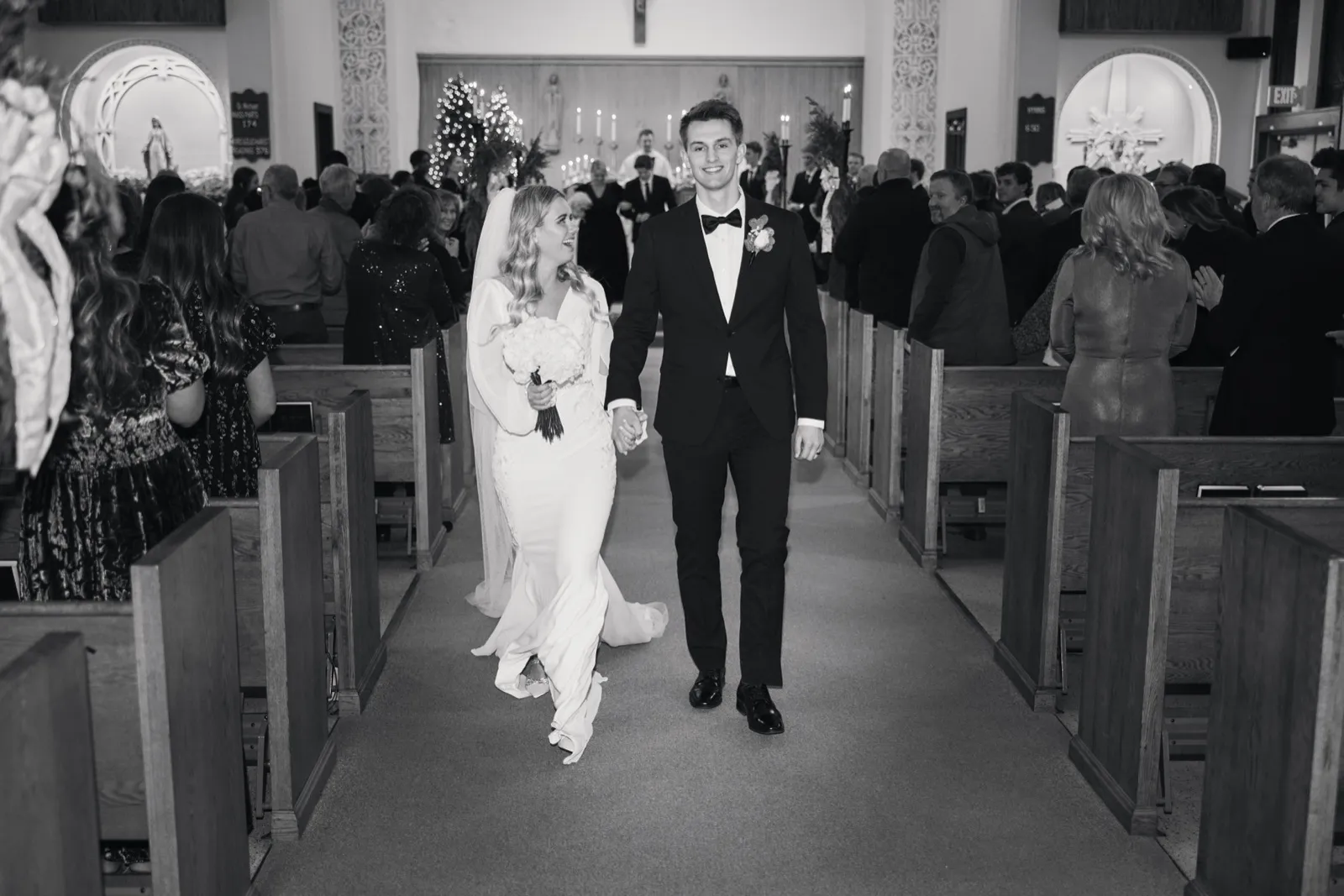 The bride and groom hold hands and walk down the church aisle after their ceremony, the bride laughing as guests stand in the pews on either side and a lit Christmas tree glows behind the altar. — Tim Larsen Photography, Brainerd Lakes MN