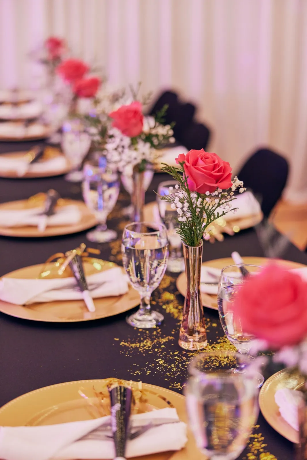A reception table set with gold chargers, white napkins, water glasses, and bud vases holding pink roses and baby's breath, with gold glitter scattered across a dark runner. — Tim Larsen Photography, Brainerd Lakes MN