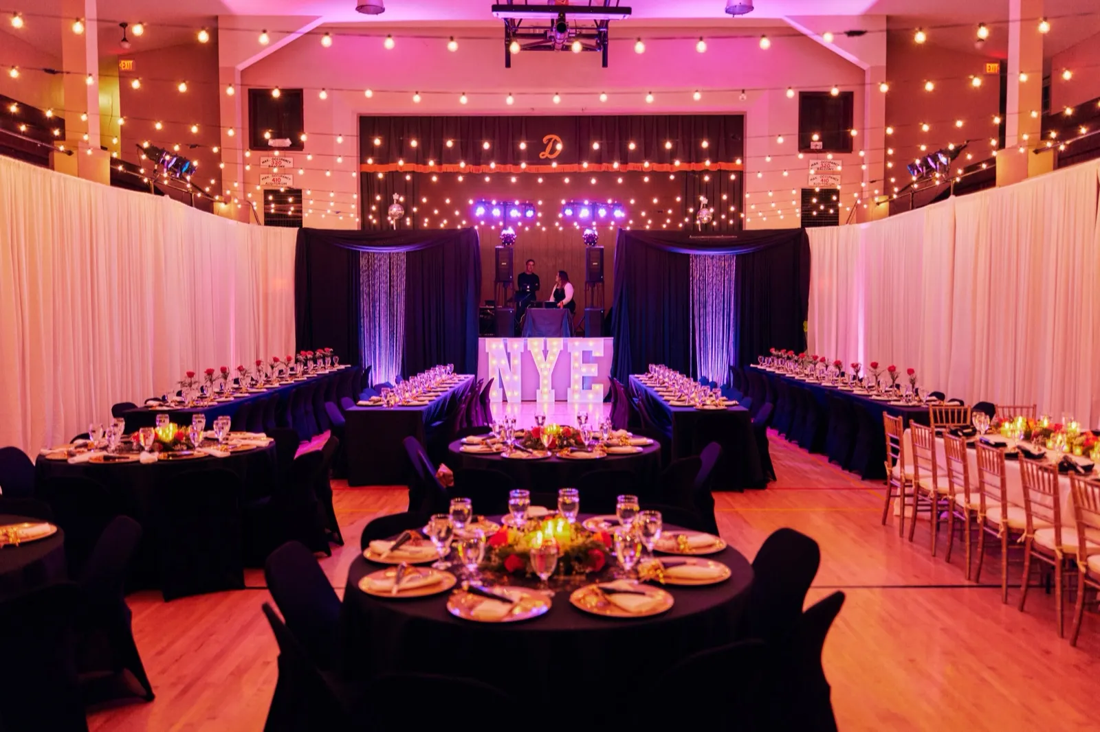 A wide view of a reception hall draped in curtains with string lights overhead, round and long tables set with red roses and candles, and a glowing NYE marquee sign in front of the DJ stage. — Tim Larsen Photography, Brainerd Lakes MN