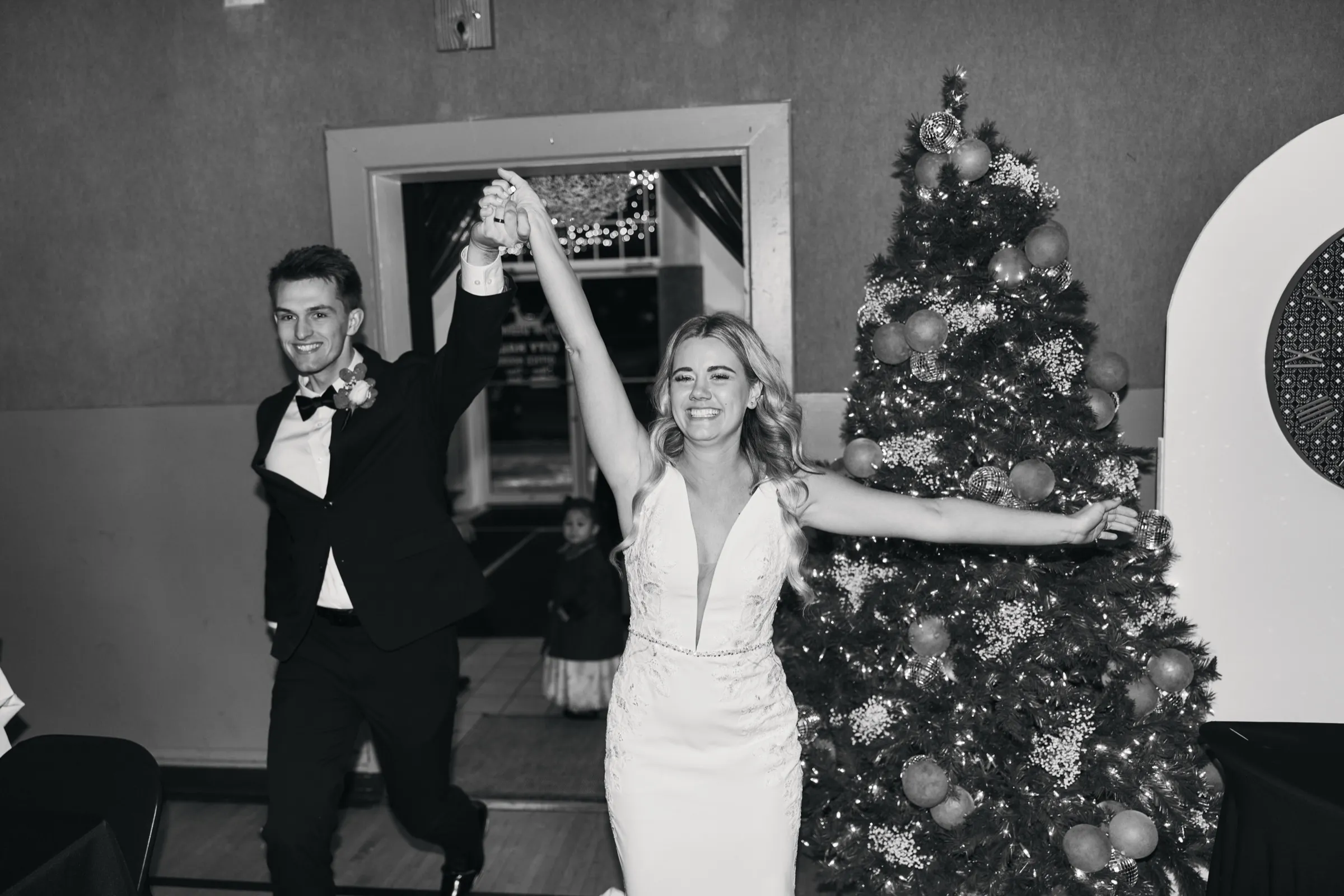 Madeline and Sam raise their joined hands in triumph as they enter the reception, a lit Christmas tree beside them and a flower girl in the doorway behind — Tim Larsen Photography, Brainerd Lakes MN