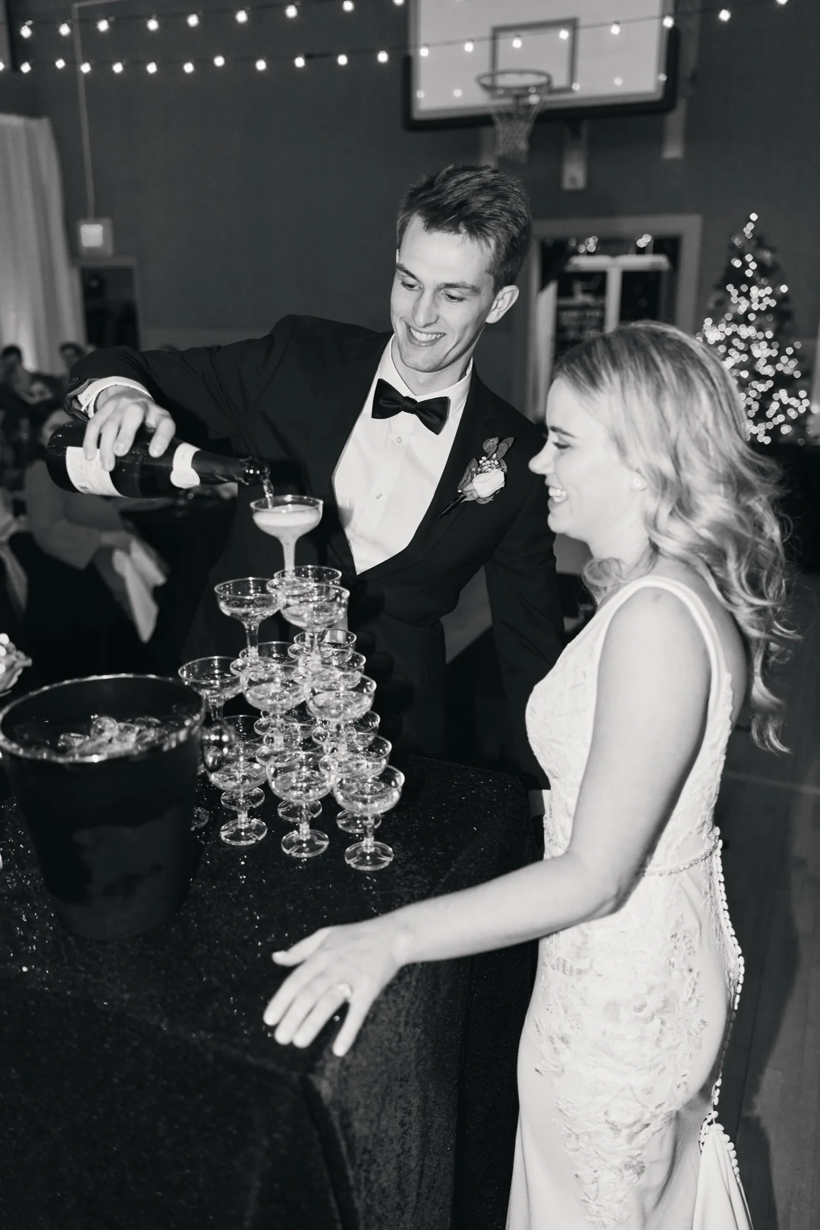 In black and white, Sam pours champagne into a tower of coupe glasses while Madeline laughs beside him, a Christmas tree and string lights behind them — Tim Larsen Photography, Brainerd Lakes MN