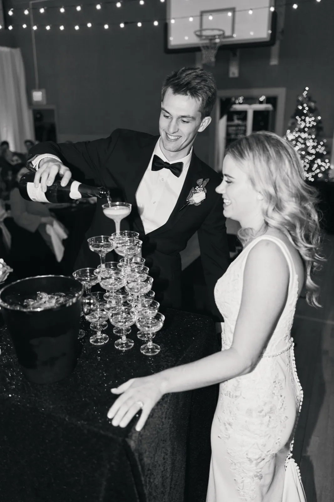 In black and white, the groom pours champagne into a tower of coupe glasses while the bride laughs beside him, with a Christmas tree and string lights in the background. — Tim Larsen Photography, Brainerd Lakes MN