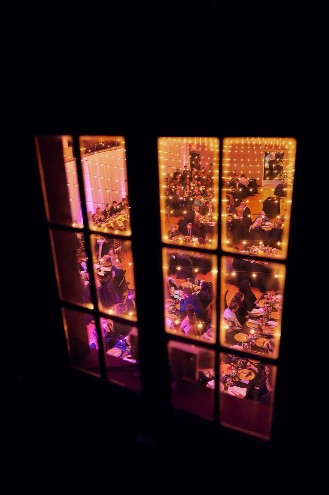 A wedding reception glows warmly through the panes of a darkened exterior window at night, with guests dining beneath strings of fairy lights inside. — Tim Larsen Photography, Brainerd Lakes MN