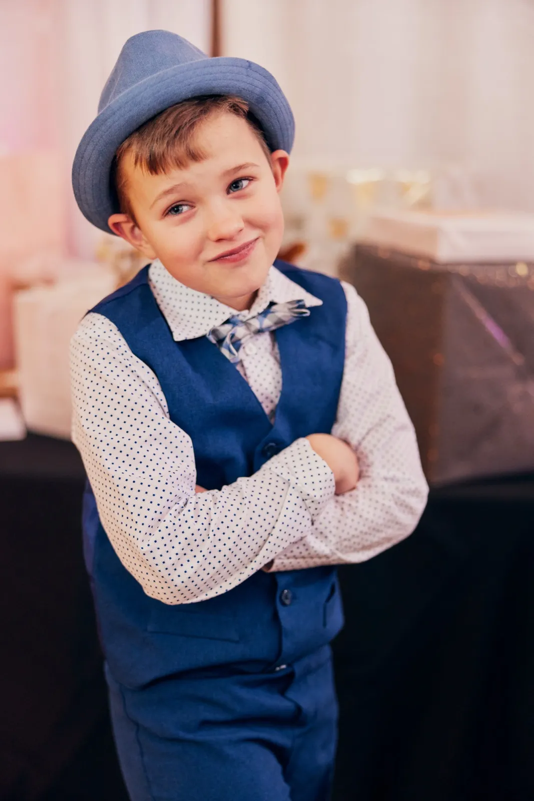 A young boy in a blue fedora, polka-dot shirt, plaid bow tie, and blue vest stands with arms crossed, smirking slightly at the camera in a softly lit reception space. — Tim Larsen Photography, Brainerd Lakes MN