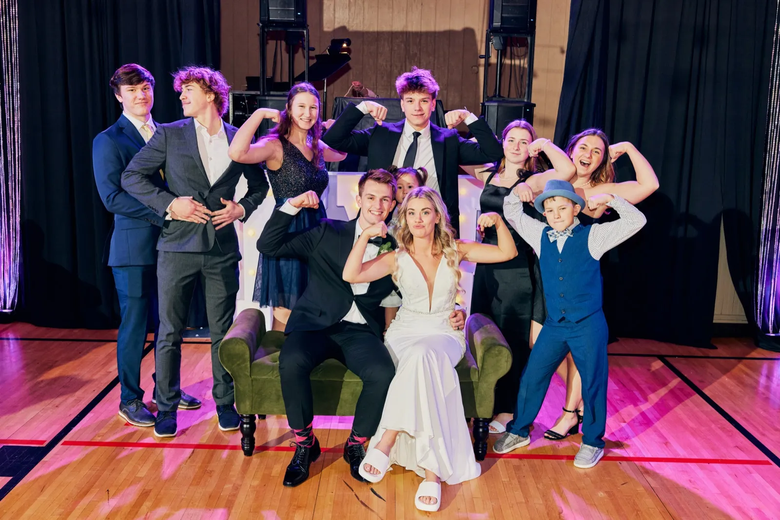 The bride sits on a green velvet sofa flanked by the groom and eight bridal-party members, most flexing their arms playfully toward the camera at an indoor reception. — Tim Larsen Photography, Brainerd Lakes MN
