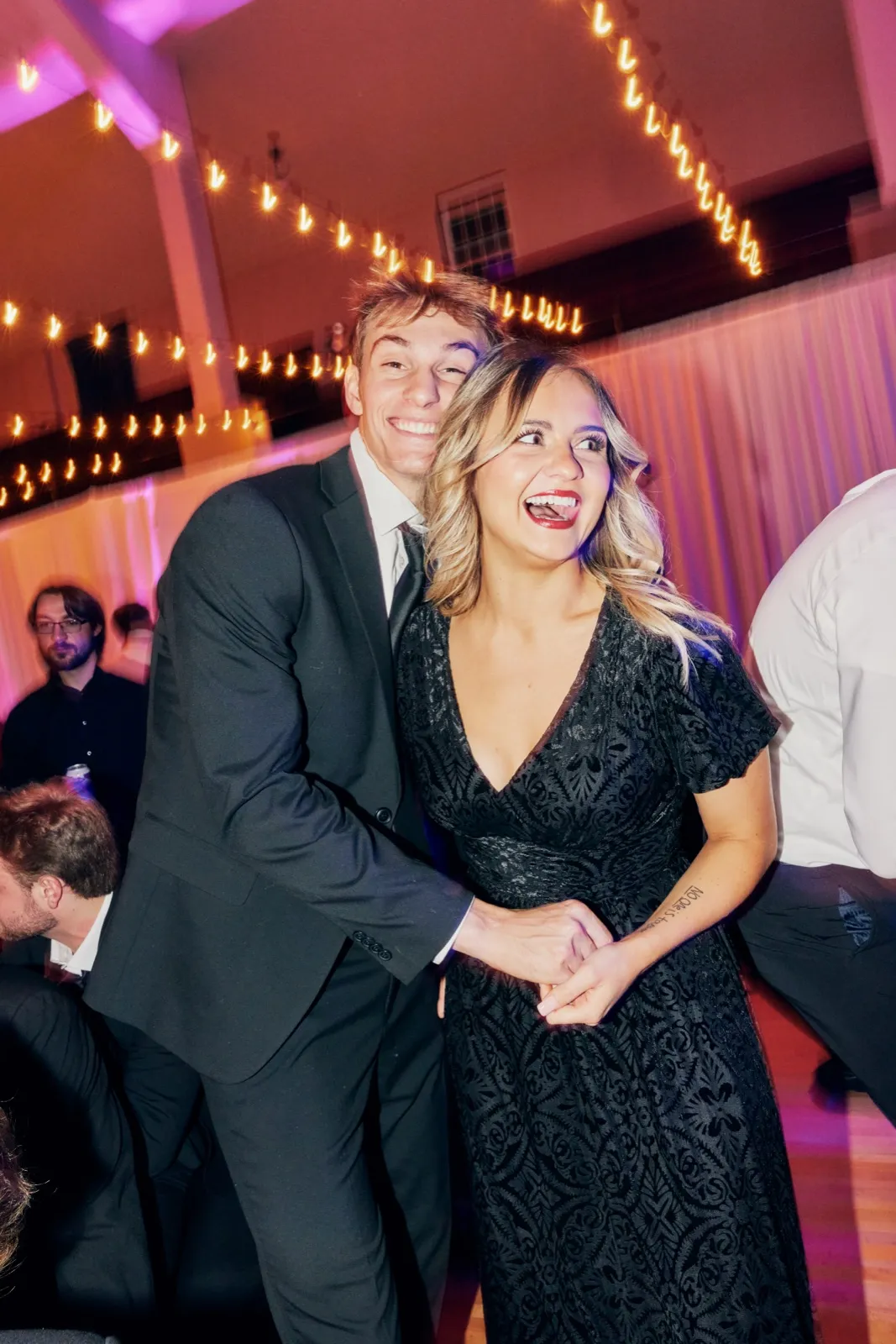 A young man in a black suit and a woman in a black patterned dress lean cheek-to-cheek, both laughing under canopies of warm string lights on a reception dance floor. — Tim Larsen Photography, Brainerd Lakes MN