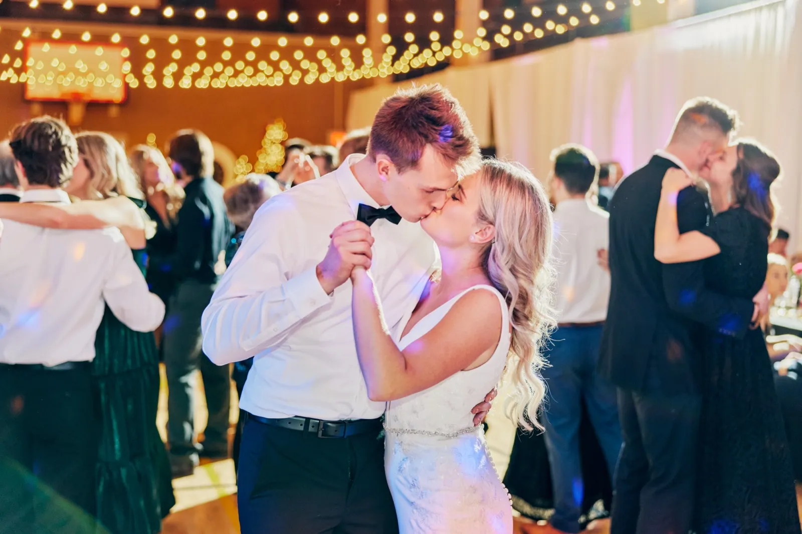 The bride and groom share a kiss on the dance floor surrounded by other dancing couples beneath a canopy of warm string lights. — Tim Larsen Photography, Brainerd Lakes MN