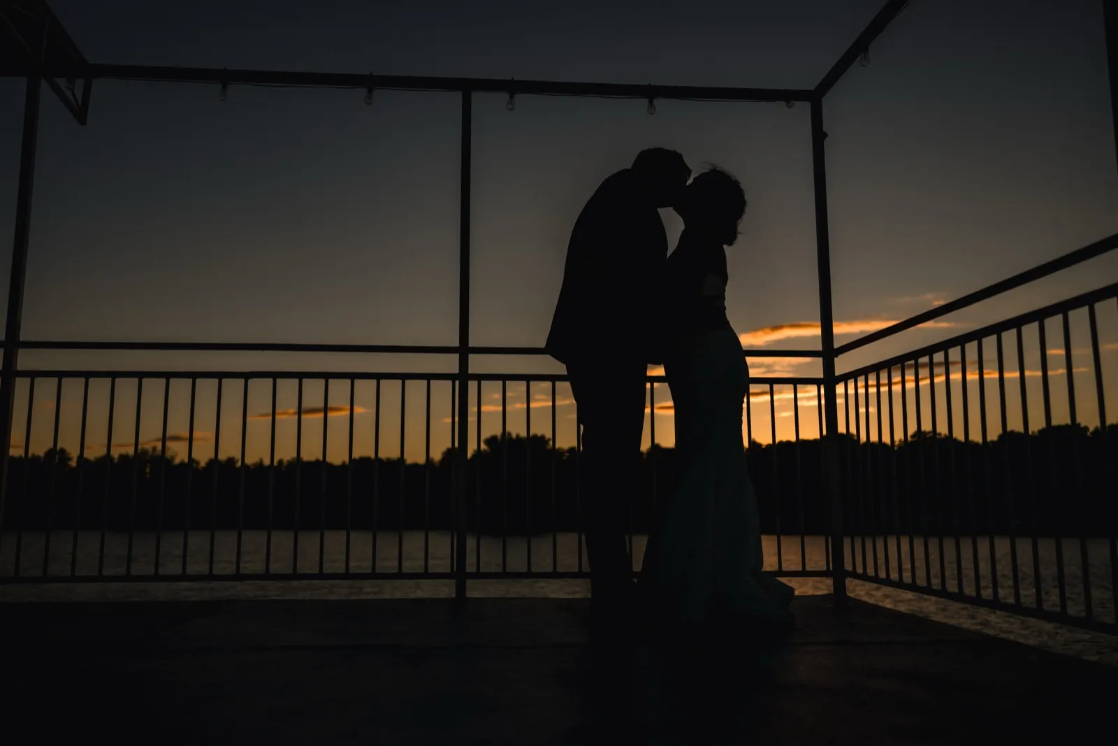 The bride and groom kiss in silhouette beneath a string-lit dock pavilion as the sky burns orange over the lake at dusk — Tim Larsen Photography, Brainerd Lakes MN