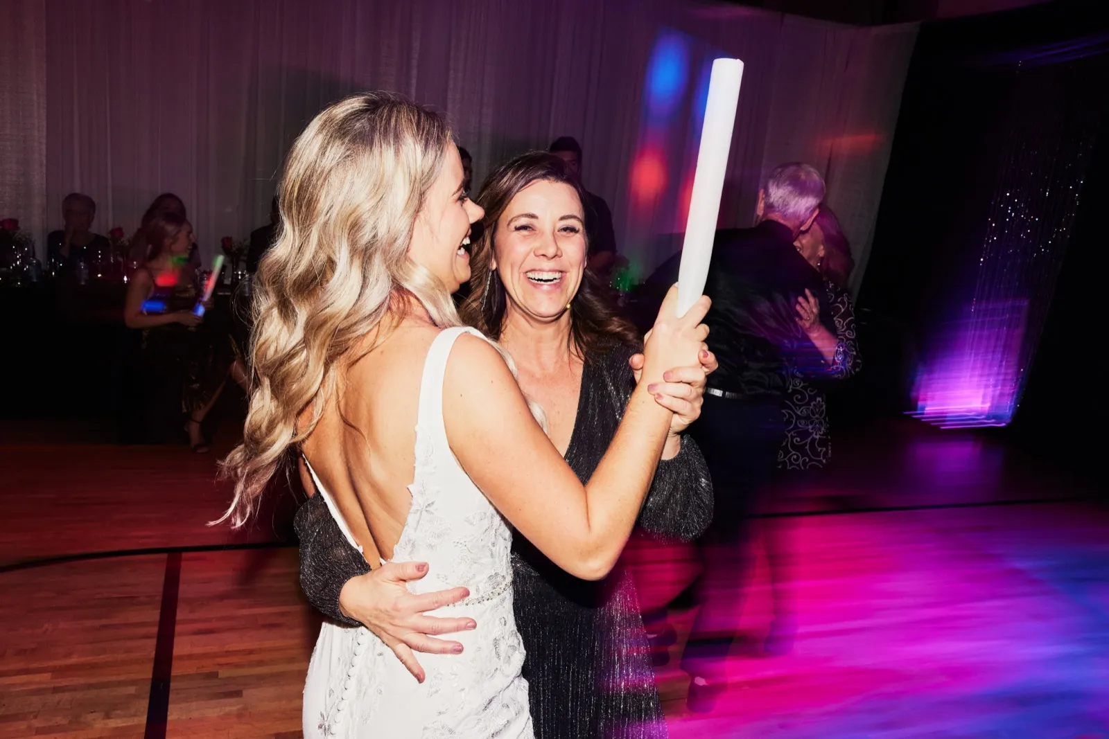 The bride laughs face-to-face with a guest who holds a glowing foam stick aloft on a magenta-lit dance floor. — Tim Larsen Photography, Brainerd Lakes MN