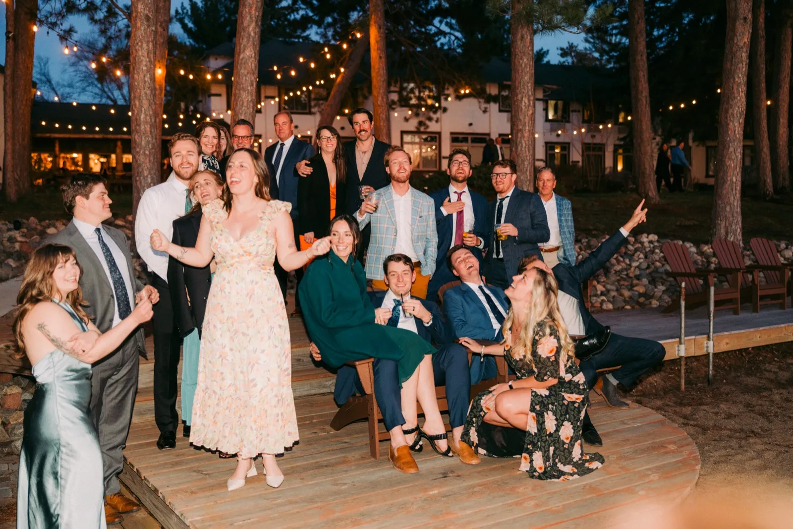 The bride dances and laughs on a wooden deck surrounded by the wedding party and family at dusk, with string lights and the lodge glowing behind them — Tim Larsen Photography, Brainerd Lakes MN