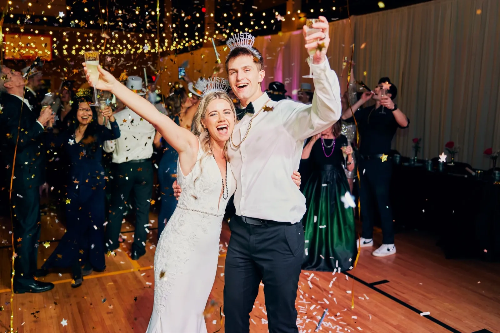 The bride and groom raise champagne glasses and cheer as gold confetti falls around them on a dance floor, with guests in tiaras and party hats celebrating behind them. — Tim Larsen Photography, Brainerd Lakes MN