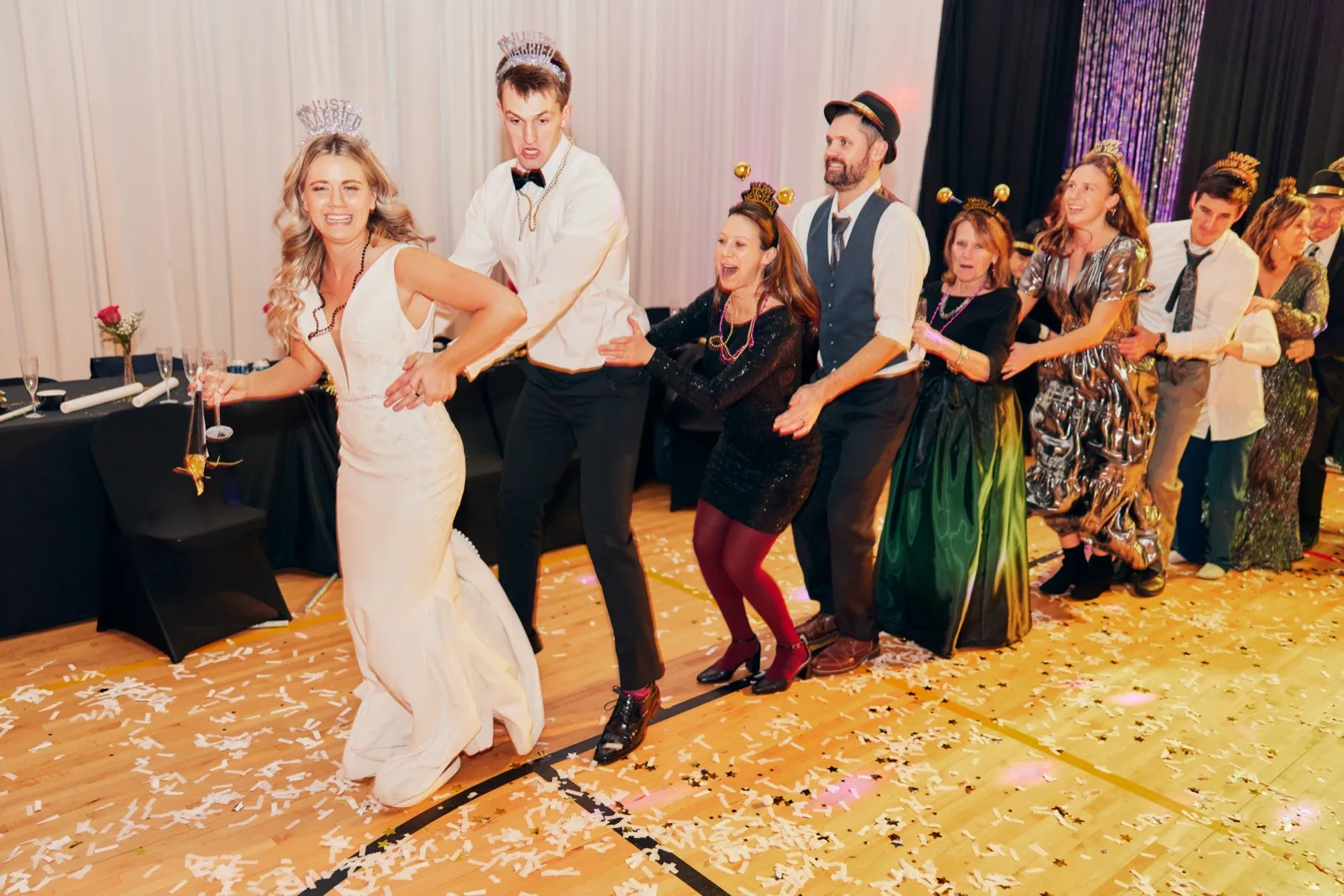 The bride leads a conga line of guests across a confetti-strewn dance floor while wearing a Just Married tiara and holding a noisemaker, with the groom and party-hatted guests following behind. — Tim Larsen Photography, Brainerd Lakes MN