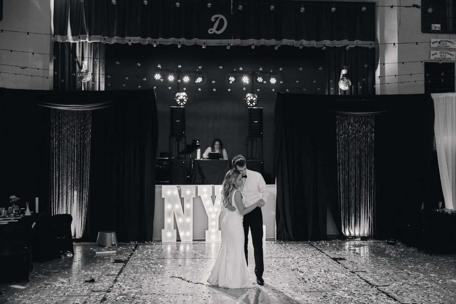 The couple share a first dance on a confetti-strewn floor in front of a stage flanked by tall draped curtains and a lit marquee sign. — Tim Larsen Photography, Brainerd Lakes MN