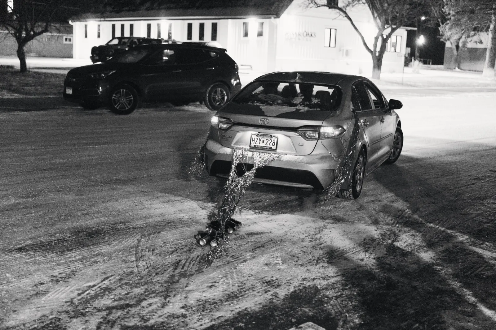 A sedan in a snowy parking lot at night with cans and streamers tied to its bumper, trailing across the icy ground. — Tim Larsen Photography, Brainerd Lakes MN