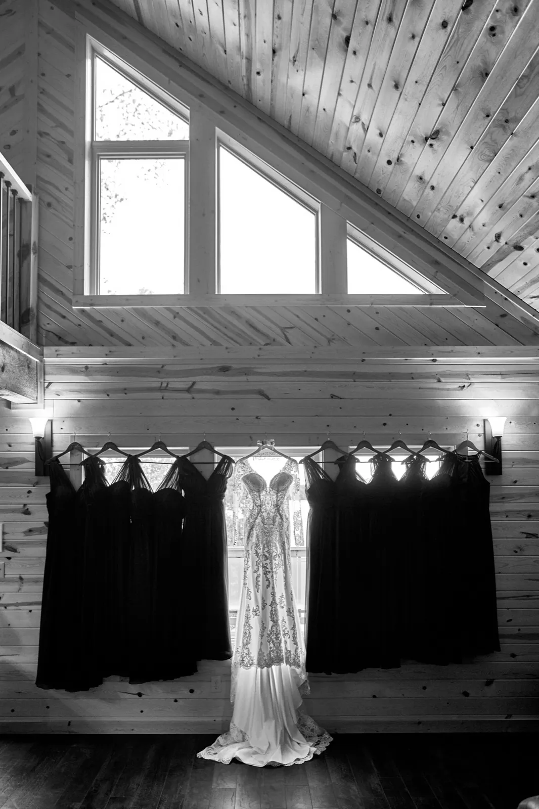 The bride's lace gown hangs at the center of a tall cabin window, flanked by bridesmaid dresses (black and white) — Tim Larsen Photography, Brainerd Lakes MN