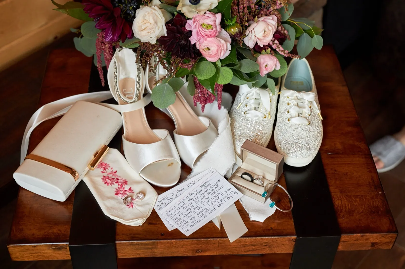 A wedding-day flat lay — jewel-toned bouquet, heels and glitter sneakers, the ring, an embroidered handkerchief, and the bride's handwritten vows — Tim Larsen Photography, Brainerd Lakes MN