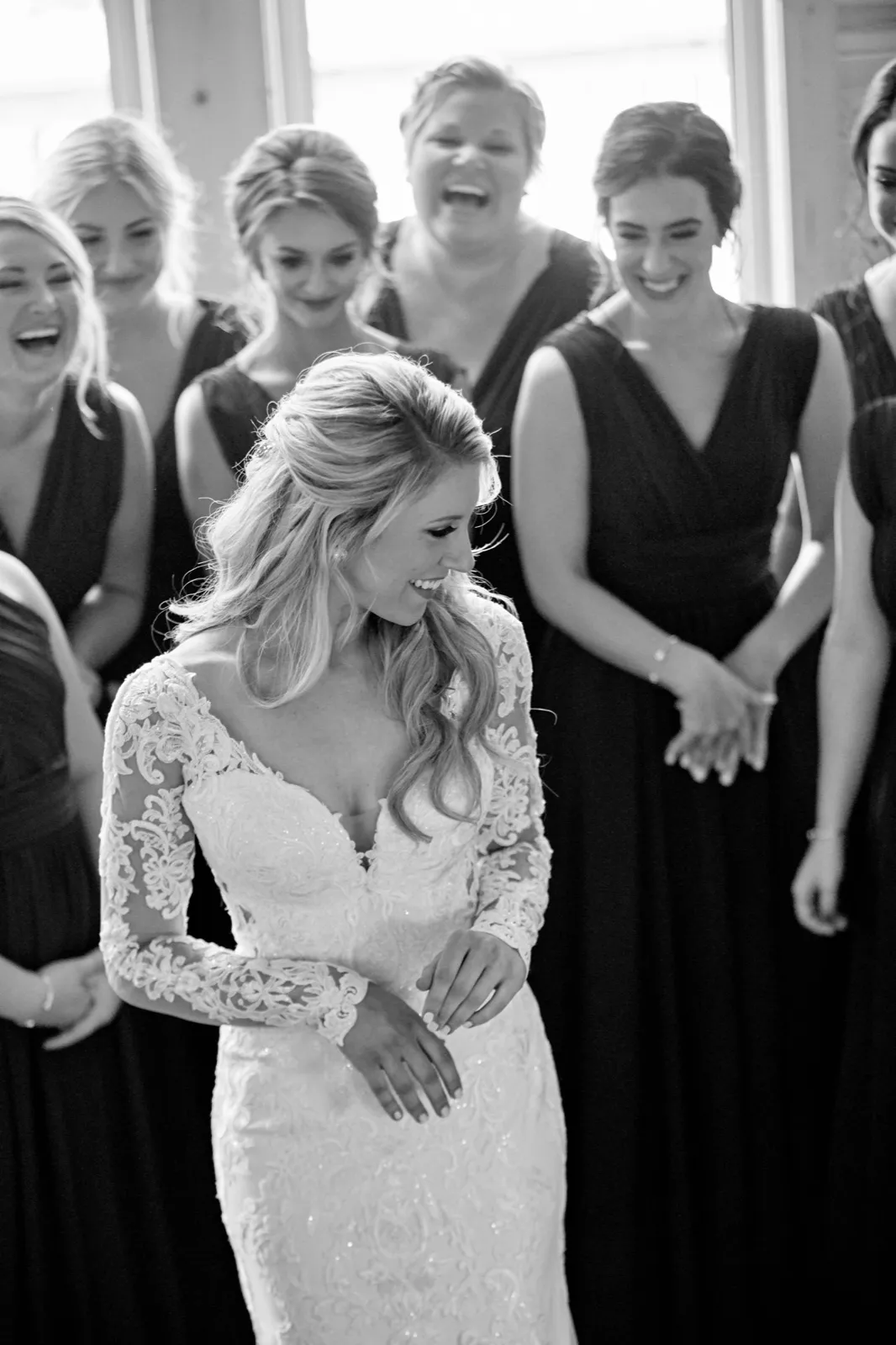 The bride laughs looking down at her lace gown as her bridesmaids react with joy behind her in a window-lit room — Tim Larsen Photography, Brainerd Lakes MN