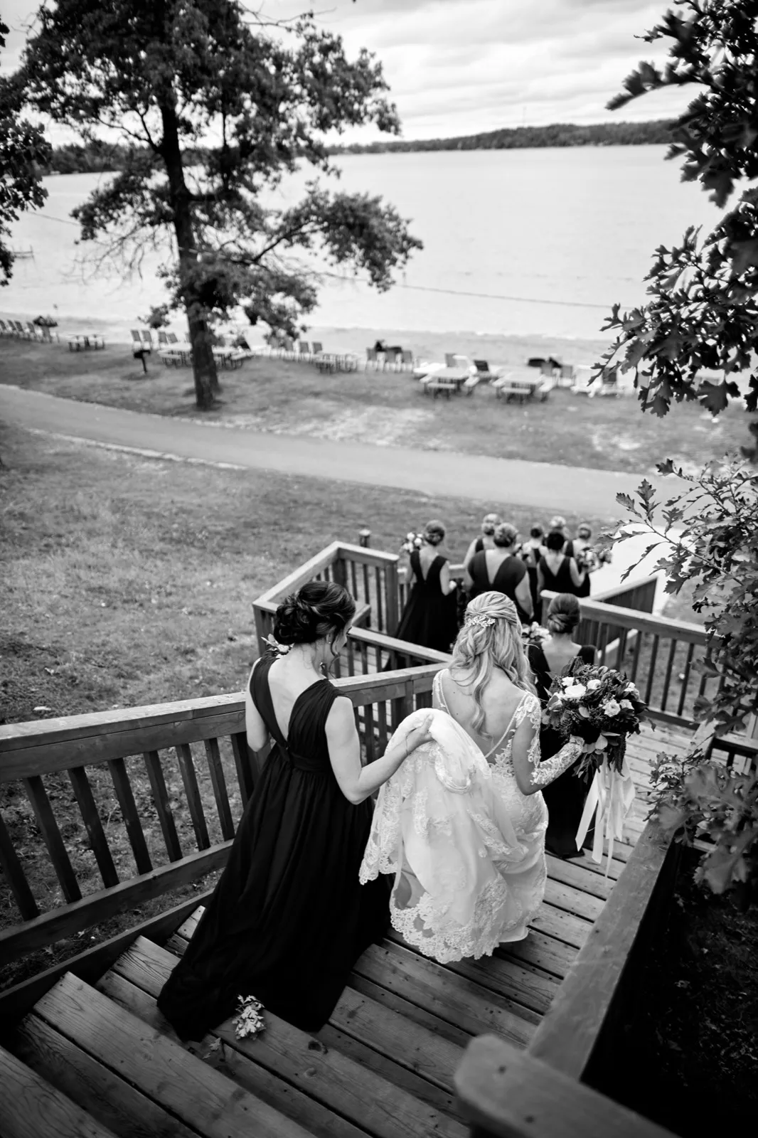 From above, the bride descends a wooden staircase with a bridesmaid lifting her train, Gull Lake stretching out behind at Cragun's Resort — Tim Larsen Photography, Brainerd Lakes MN