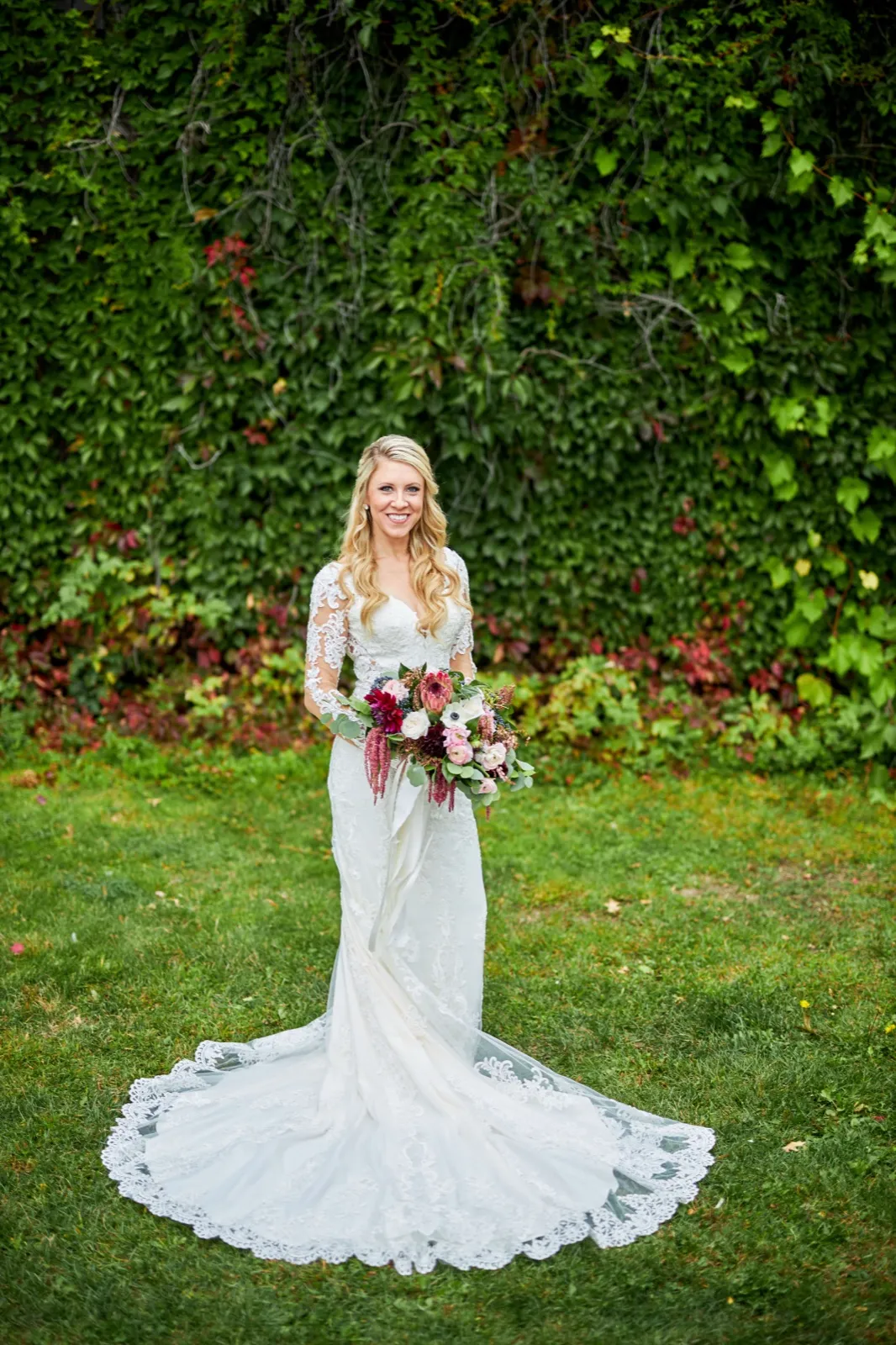 The bride stands on a lawn holding a burgundy and blush bouquet, her lace train fanned out against a tall ivy wall — Tim Larsen Photography, Brainerd Lakes MN
