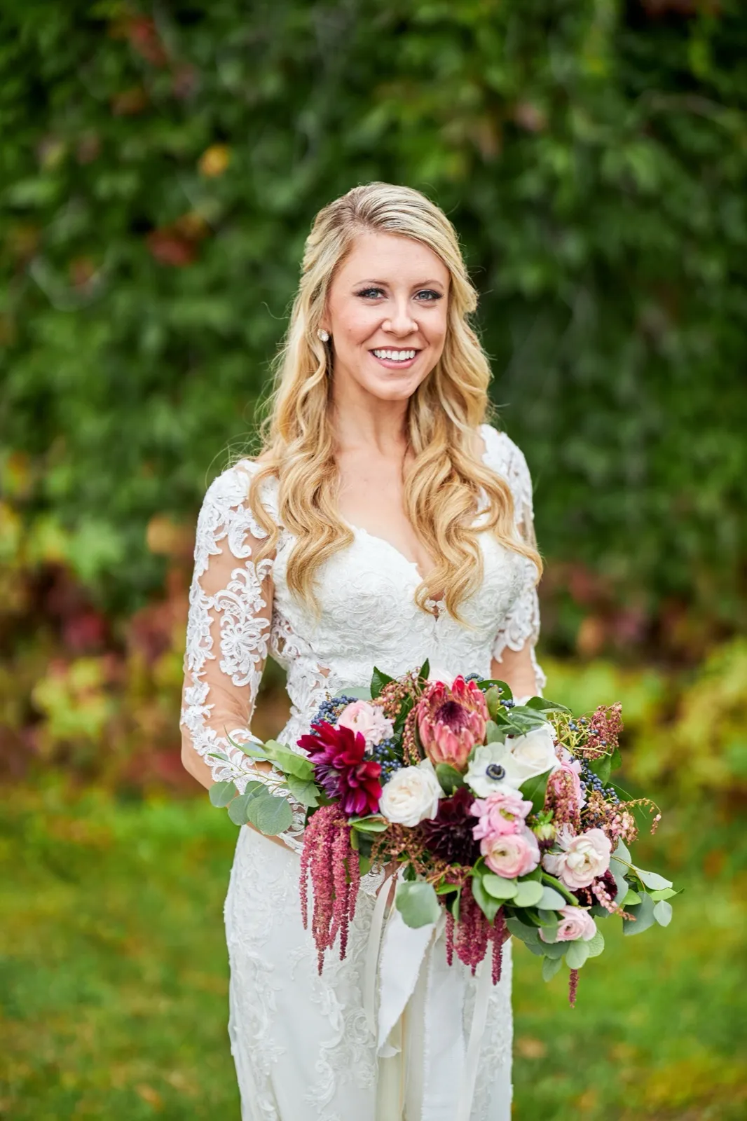 The bride smiles holding a large cascading bouquet of jewel-toned florals against green foliage — Tim Larsen Photography, Brainerd Lakes MN
