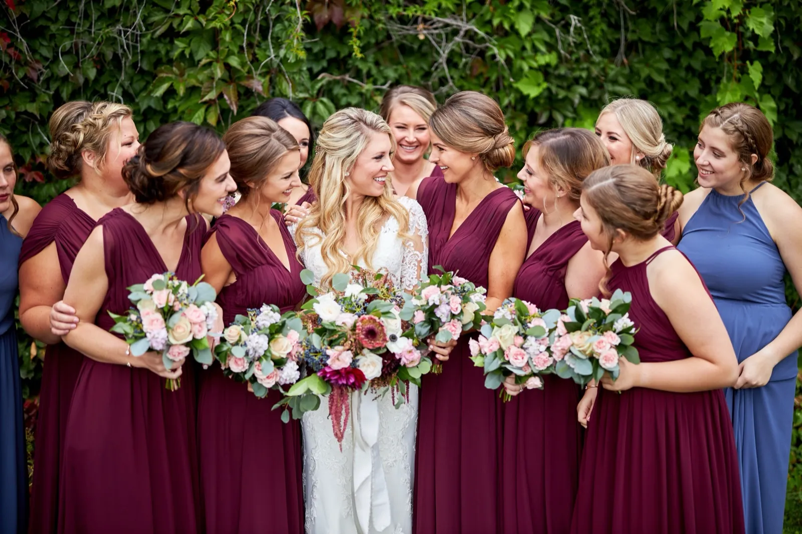 The bride at the center of her bridesmaids in burgundy and blue, all laughing together against an ivy-covered wall — Tim Larsen Photography, Brainerd Lakes MN