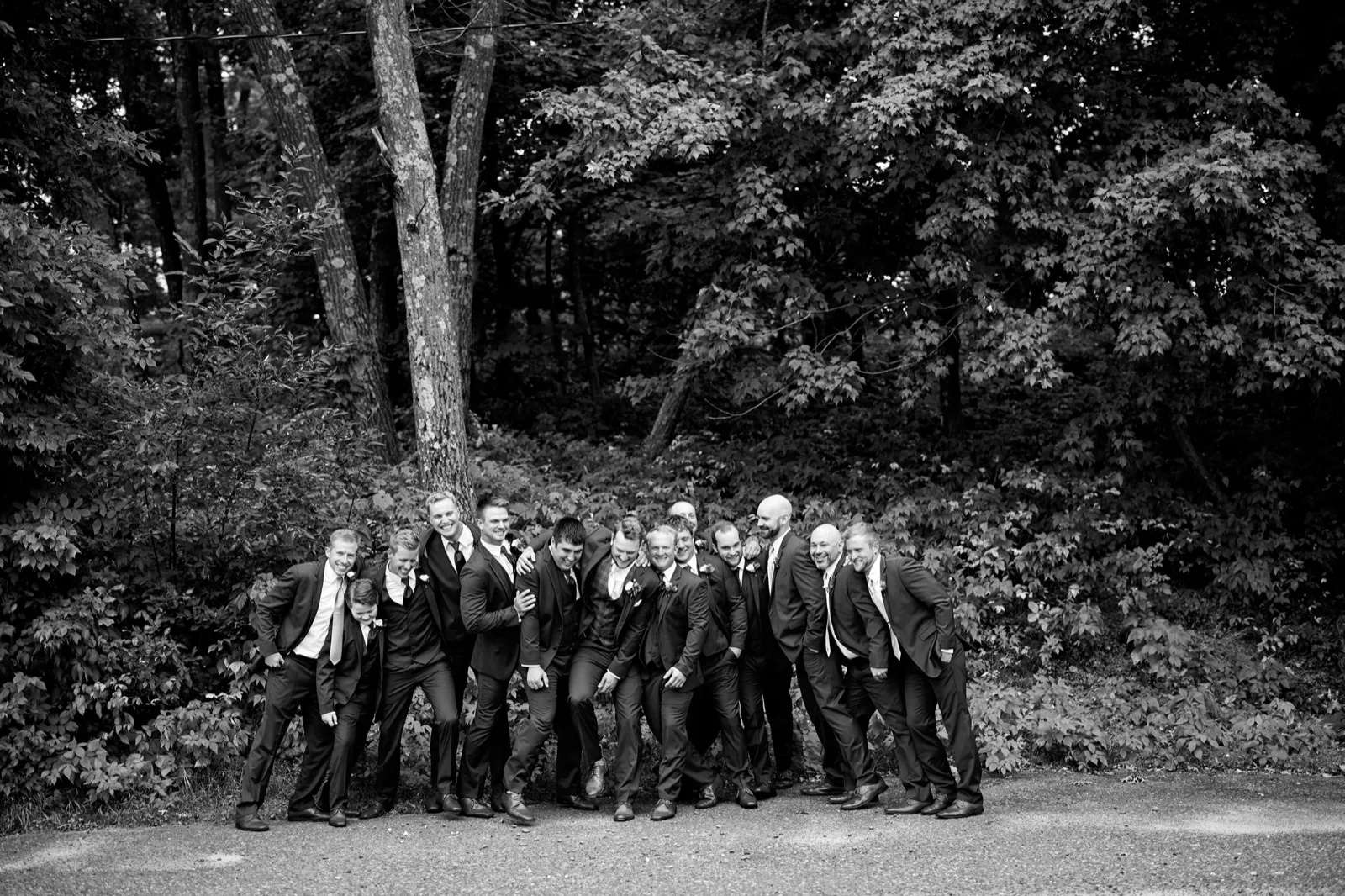 The groom and his groomsmen lean together laughing on a wooded road (black and white) — Tim Larsen Photography, Brainerd Lakes MN
