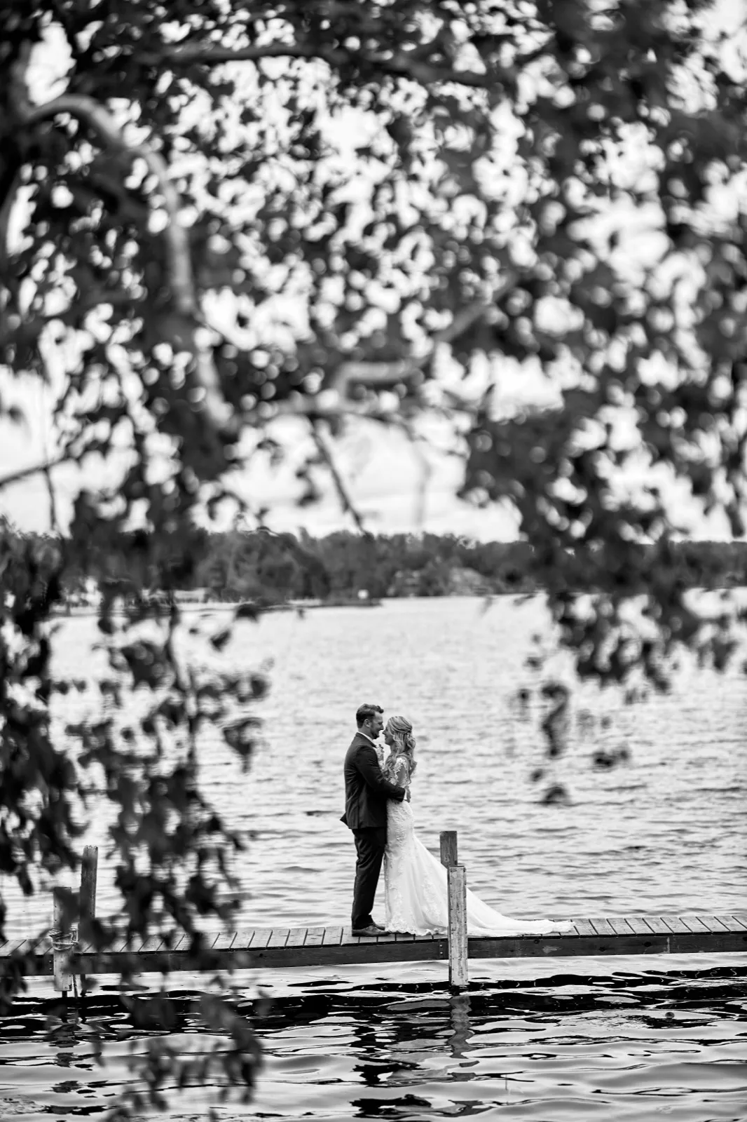 The bride and groom stand forehead-to-forehead on a dock over the lake, framed through out-of-focus leaves — Tim Larsen Photography, Brainerd Lakes MN