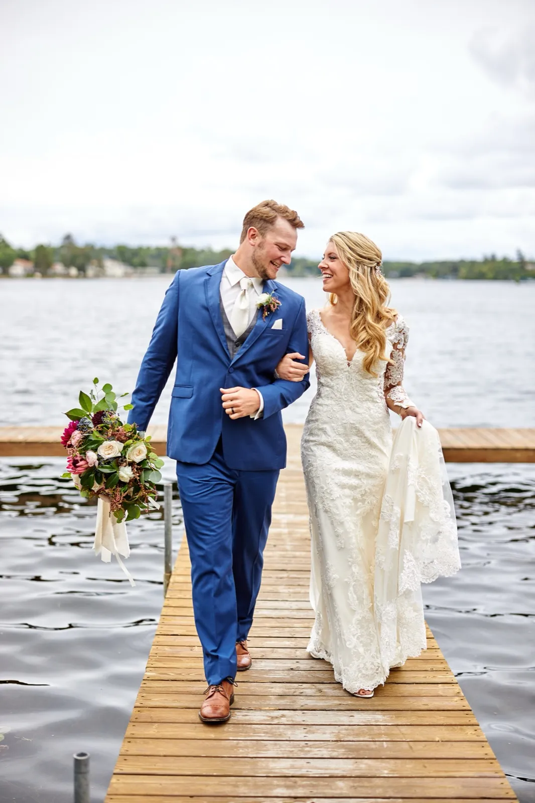 The bride and groom walk down a dock laughing, the bride holding her dress and a trailing bouquet of garden roses — Tim Larsen Photography, Brainerd Lakes MN