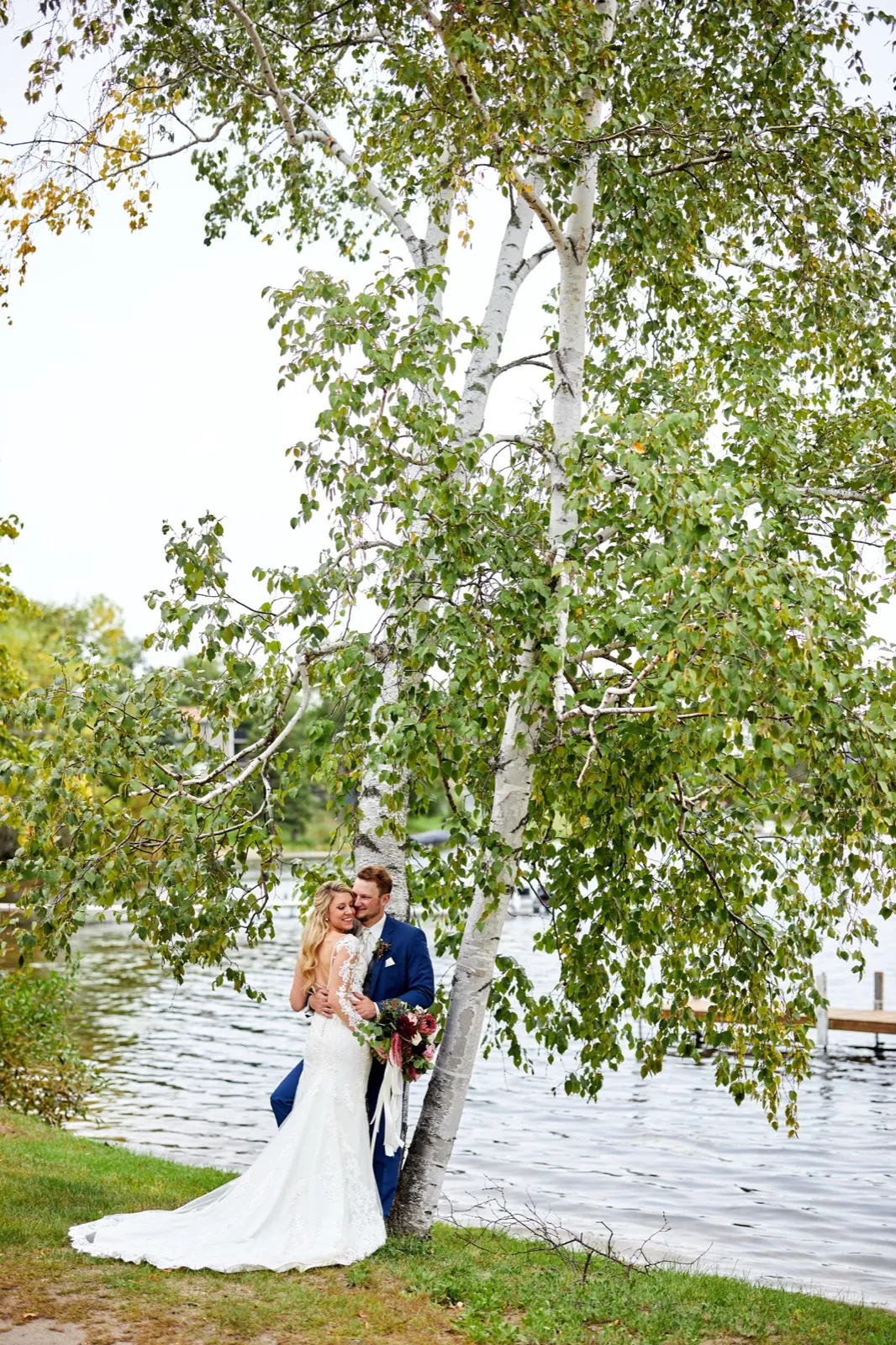 The bride and groom embrace at the base of a birch on the Gull Lake shore, the dock behind them at Cragun's Resort — Tim Larsen Photography, Brainerd Lakes MN