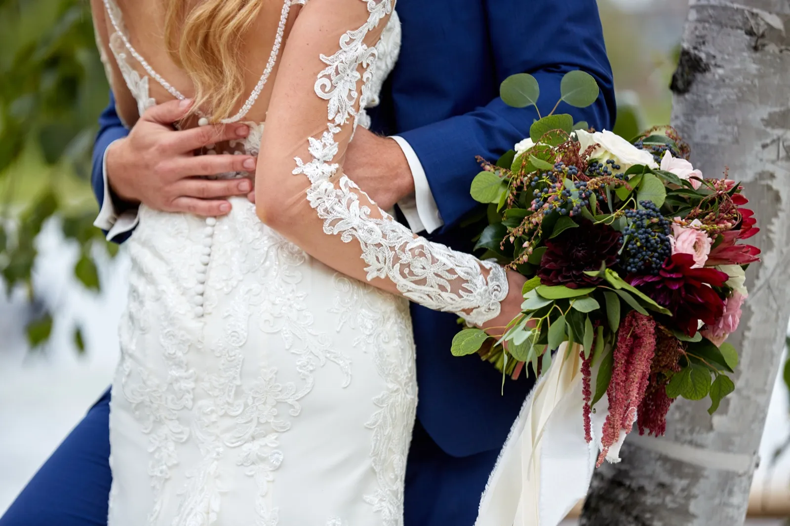Close-up of the groom's hand on the bride's lace back as she holds a burgundy and blush bouquet beside a birch — Tim Larsen Photography, Brainerd Lakes MN