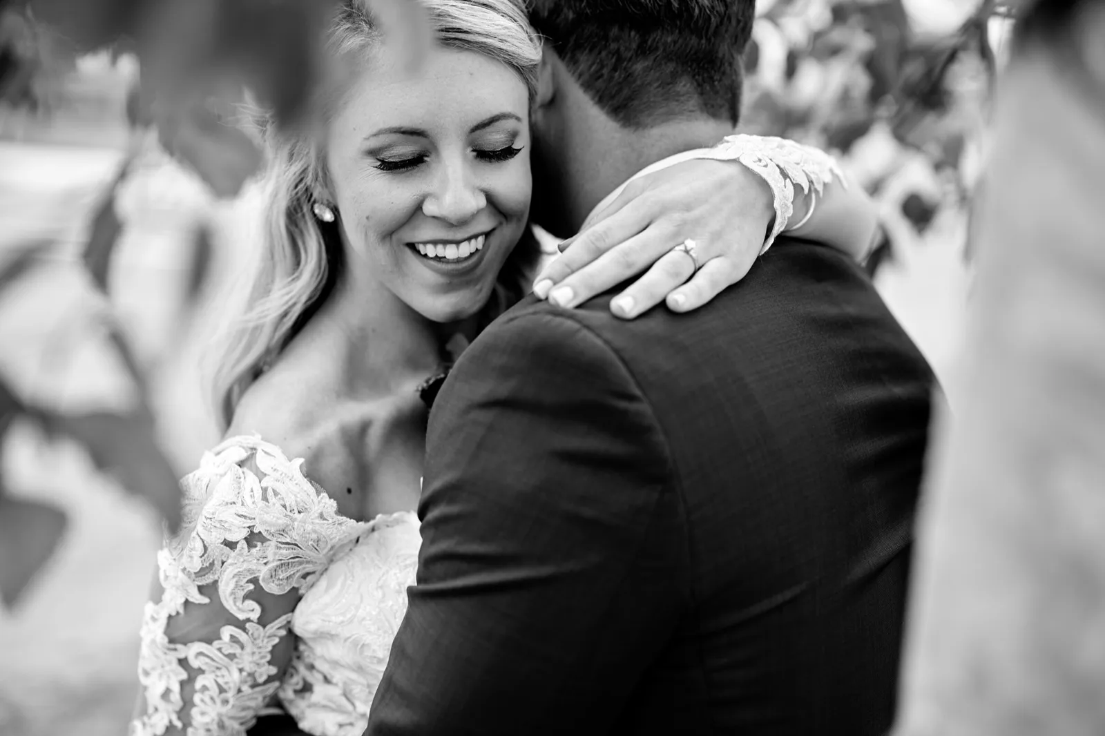 The bride smiles with her eyes closed against the groom's shoulder, her ringed hand on his jacket, soft foliage around them — Tim Larsen Photography, Brainerd Lakes MN