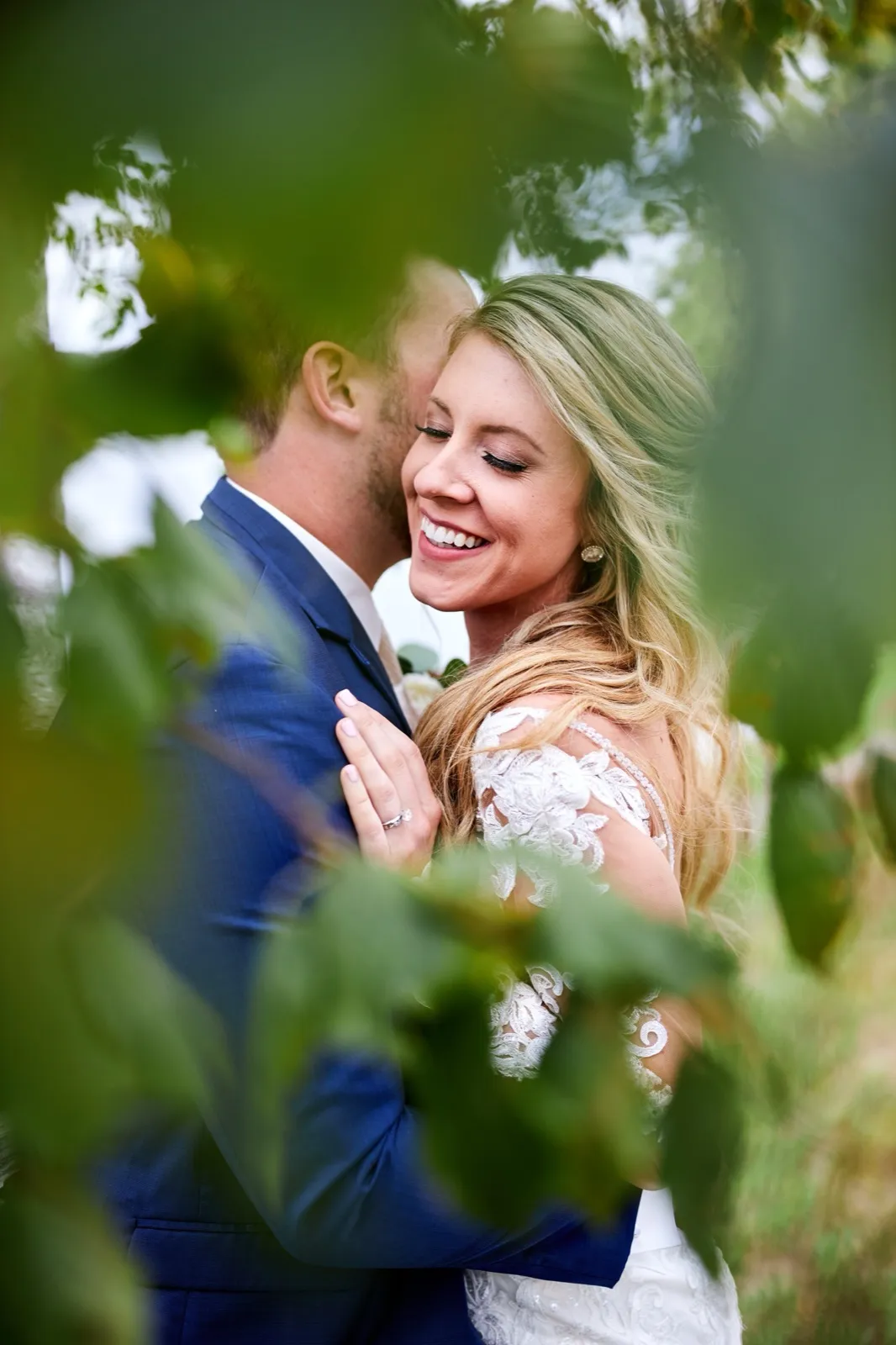 The bride smiles with closed eyes against the groom's cheek as they embrace, seen through a frame of green leaves — Tim Larsen Photography, Brainerd Lakes MN