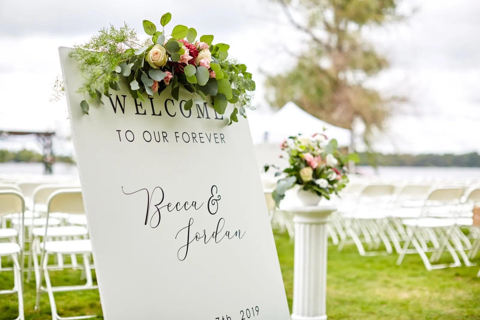 A 'Welcome to our forever, Becca & Jordan' sign topped with florals beside rows of white chairs on the lakeside ceremony lawn — Tim Larsen Photography, Brainerd Lakes MN
